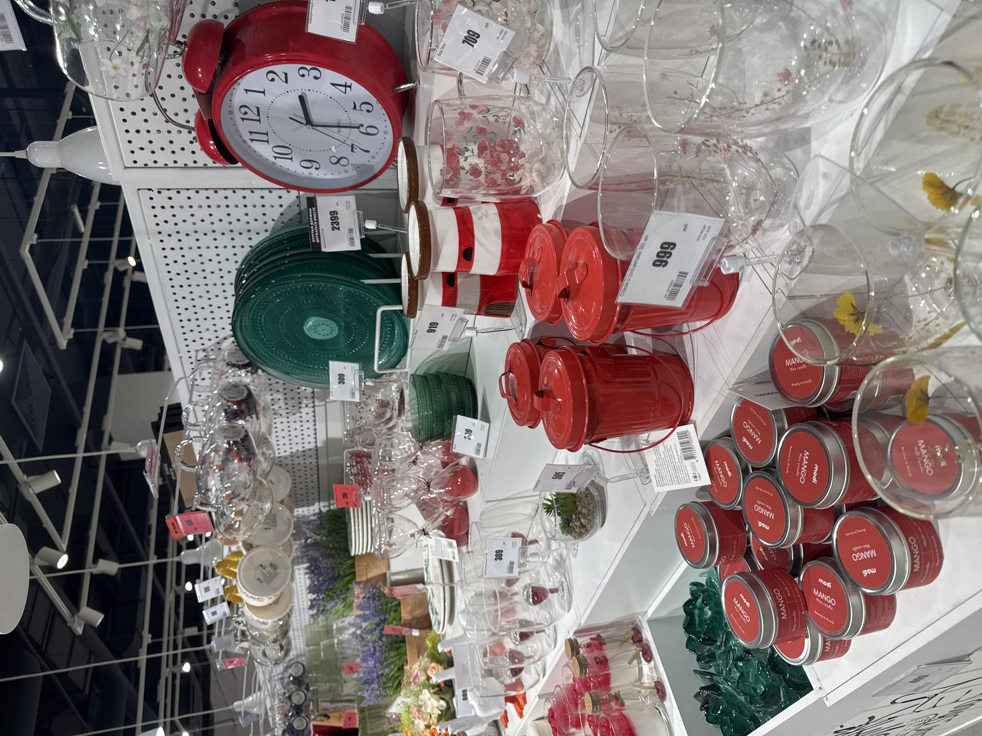The image depicts a retail display of household goods, likely within a store. The foreground is dominated by rows of small, red, cylindrical containers, some with silver lids, stacked and arranged on shelves. These containers are labeled "modi MANGO wax candle." To the left and slightly behind these, there are more green decorative items resembling lotus flowers and some clear glass vials with red contents and cork stoppers.
Further back, a collection of red metal trash cans is visible. To the right, clear glass tumblers with delicate floral patterns are displayed. Several decorative lighthouse-shaped candle holders in red and white are also present. Behind these, a large red alarm clock with a white face and black numbers is prominently displayed on a shelf.
In the background, various glassware, including wine glasses and stacks of plates, are visible. Potted plants, possibly lavender, add a touch of greenery to the scene. The overall environment appears to be a well-lit store with white shelving and a black ceiling with spotlights. There are price tags on many of the items. The image does not contain any people.
The specific location in Ufa, Russia, cannot be determined from the image alone.