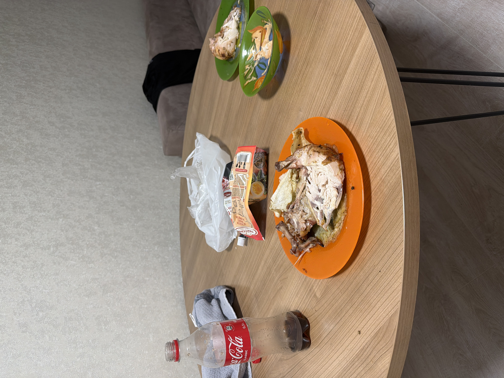 The image shows a close-up of a round wooden table with food and a drink on it. There are two plates with pieces of chicken, one orange and one green. A small green bowl with a painted image is also on the table. Next to the chicken, there is a tube of sauce. A partially empty bottle of Coca-Cola Classic sits on the table with a grey cloth behind it. A crumpled white plastic bag is also visible on the table. In the background, a sofa and a wall with textured wallpaper can be seen. The floor appears to be light-colored wood laminate. The overall scene suggests a meal has recently been consumed. This is a still life photograph with no people present. It is not possible to determine the location within the city of Barnaul, Russia from this image.