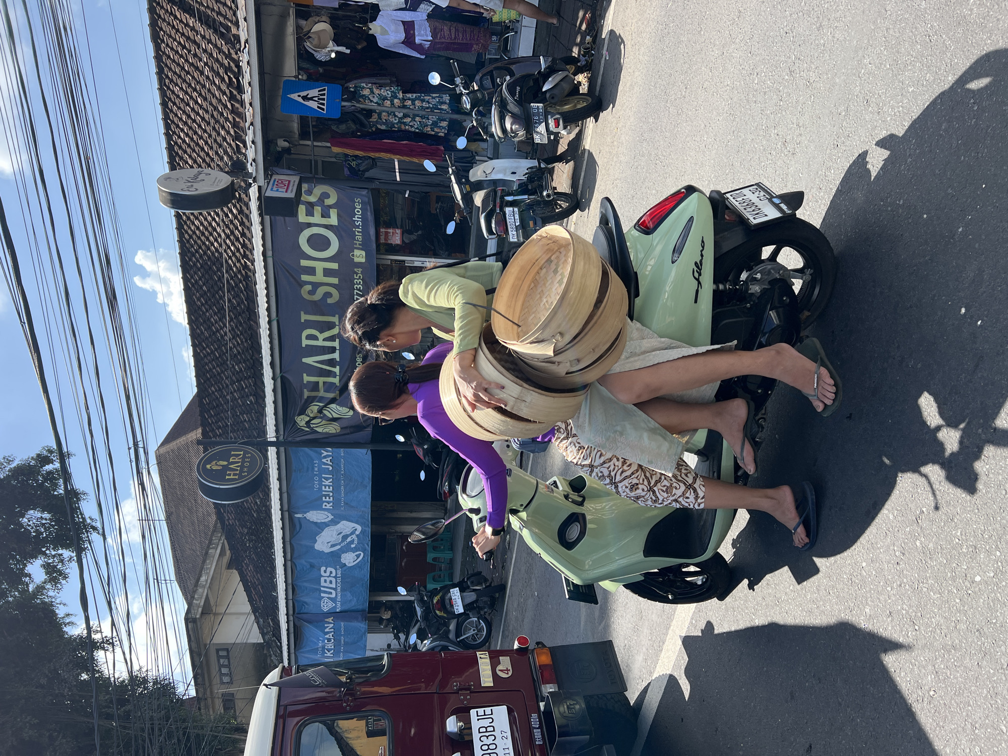Two women are riding on a light green scooter on a paved street. The woman in the front is driving and is wearing a purple long-sleeved shirt and patterned pants. The woman in the back is wearing a green long-sleeved shirt and a light-colored skirt. She is holding a stack of round, woven bamboo containers. The scooter is carrying them past a row of shops. One shop has a banner that reads "HARI SHOES". There are other scooters and a red truck parked on the street. Overhead, there are numerous electrical wires. The scene appears to be in Abiansemal, Indonesia, given the presence of the "HARI SHOES" store which is a known establishment in that location. The sky is blue with some clouds, and the sun is casting shadows on the street.