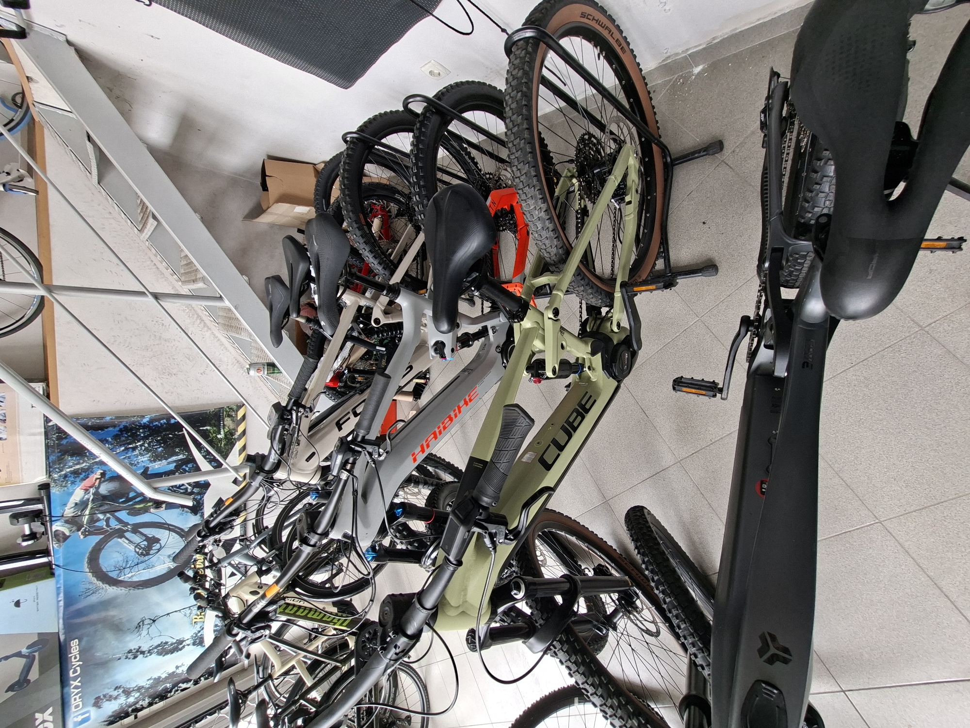The image depicts a row of bicycles, predominantly mountain bikes, stored indoors. Brands visible include Cube and Haibike. The bikes are parked in a stand, presumably in a shop or storage area. The floor is tiled. A poster featuring a mountain biker is partially visible in the background. The location is implied to be Gabrovo, Bulgaria based on the information provided.