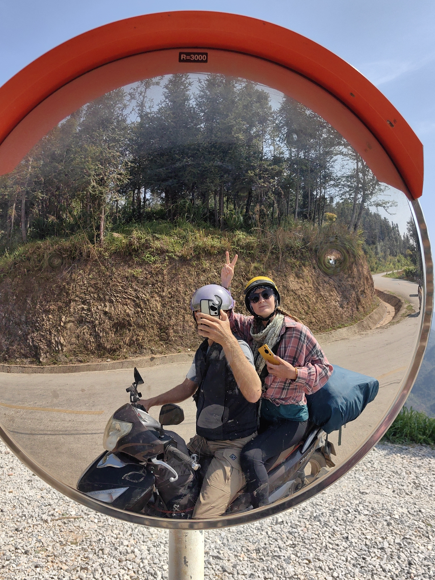 A couple is reflected in a convex mirror on a scooter in Lũng Cú Commune, Vietnam. The driver is wearing a grey helmet and a vest, holding a smartphone to take a picture. The passenger wears a yellow helmet, sunglasses, and a plaid shirt, giving a peace sign and holding a phone. A blue pack sits behind the passenger. The mirror's orange frame has "R=3000" printed on it. The background shows a tree-lined hill and a winding road. The mirror is mounted on a pole on a gravelly surface.