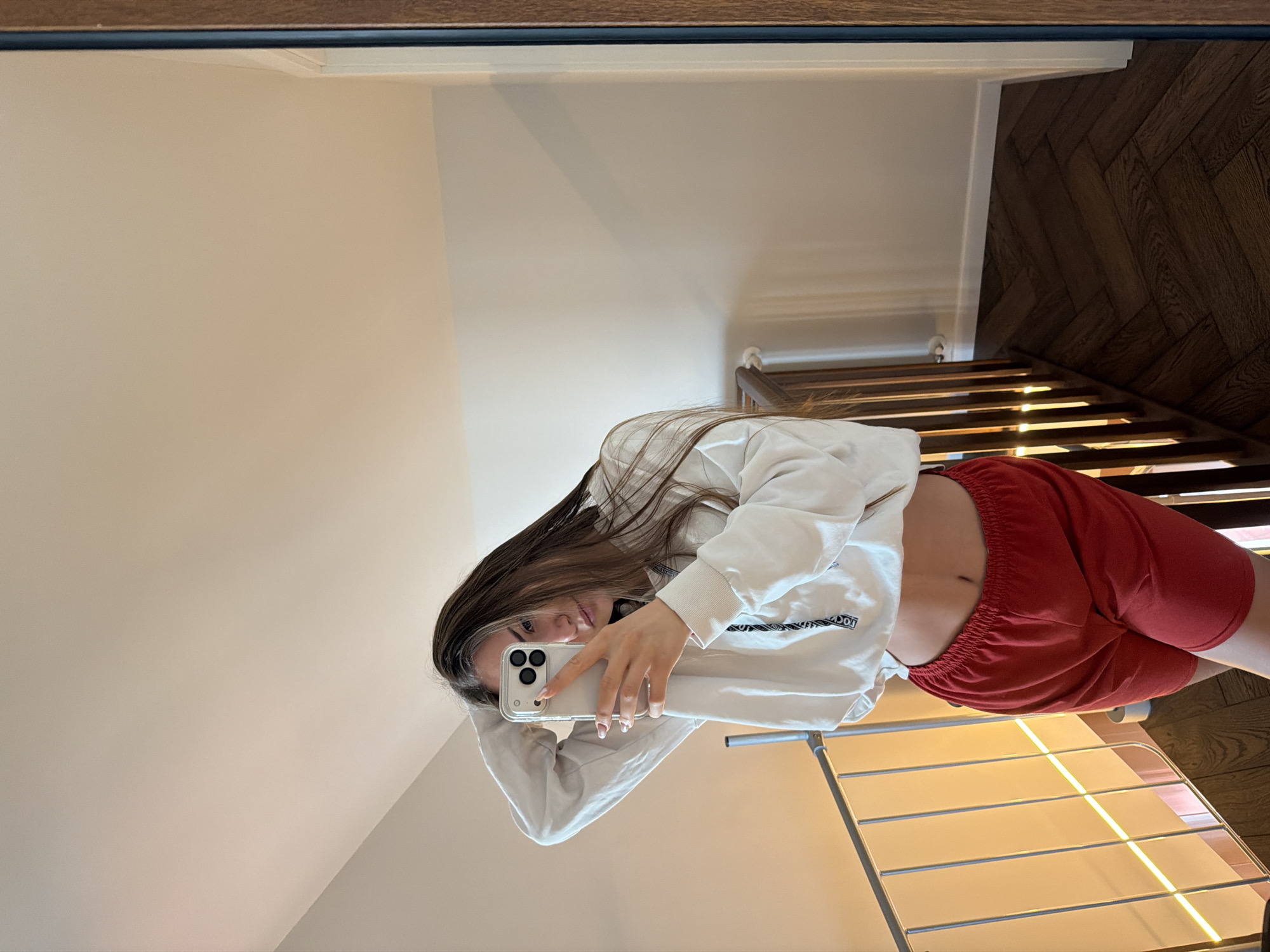 A young woman is taking a selfie in a mirror. She is wearing a white sweatshirt and red shorts, exposing her midriff. The reflection shows a room with a white wall, a wooden floor, and a metal drying rack. The location in the city of Buhovo, Bulgaria cannot be determined from the image.