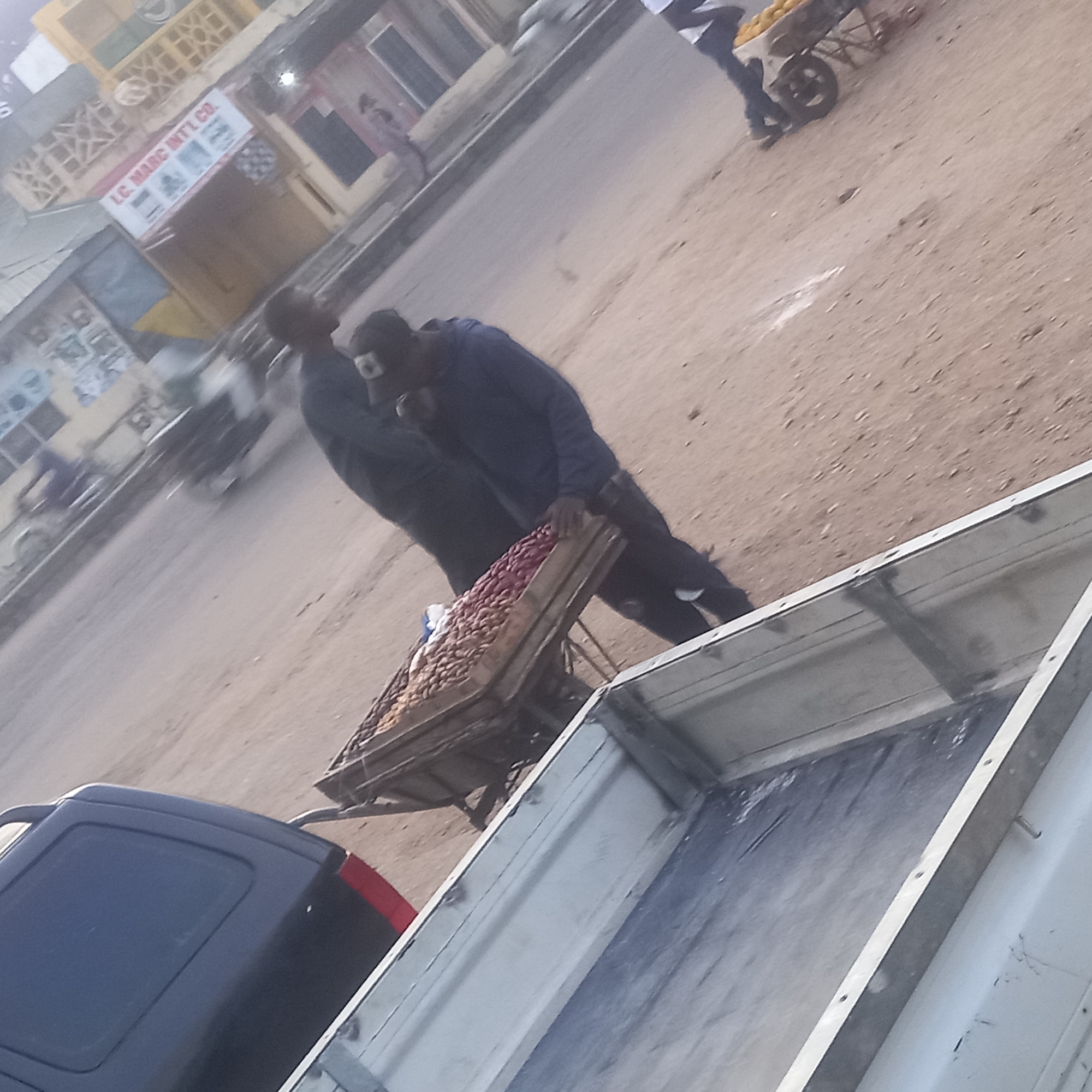 This tilted and slightly blurry image captures a bustling street scene in Dadi Riba, Nigeria, likely during the soft light of early morning or late afternoon. In the foreground, the rear of a dark blue van is partially visible. Behind it, two men in dark attire are seen actively pushing or pulling a wooden handcart. The cart is heavily laden with produce, displaying a mix of reddish-brown and lighter-colored vegetables, possibly onions or potatoes.
The road surface is unpaved, appearing dusty and sandy. Further down the street, another individual is pushing a wheelbarrow filled with bright yellow items, which appear to be fruits like mangoes. Buildings line the street in the background, suggesting commercial or residential activity. A prominent sign on one of these structures clearly displays the text "LC. MARC INTL CO." A blurred motorcycle or scooter is also visible on the road, indicating movement. The overall scene portrays a typical day of street vending and daily commerce in a local, lively environment. The activity depicts people transporting goods for sale. No specific emotions are discernible due to the distance and blur, but the scene conveys industriousness.
