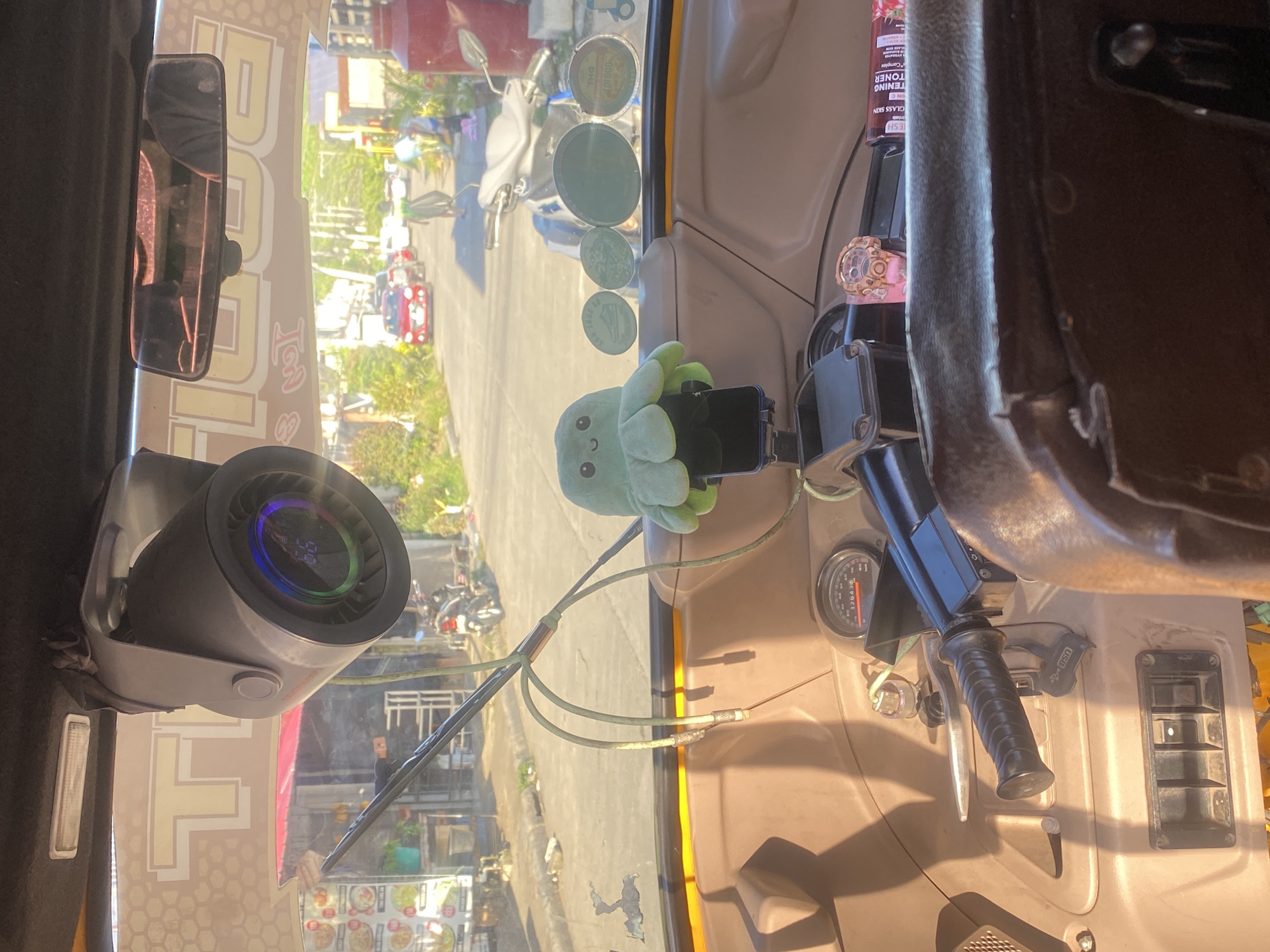 The image is a forward-facing interior view from the driver's seat of a motorized tricycle, likely in Davao City, Philippines. The scene is characterized by bright, natural lighting casting long shadows. Prominently displayed in the foreground is the steering column with a black handlebar grip and a visible speedometer showing "28438" and a needle indicating a speed between 0 and 20. To the left of the steering column is a panel with three switches and a red hazard light button. A black plastic holder for a mobile phone is mounted centrally, topped by a green, plush octopus toy with a smiling face. Above the dashboard, a circular device with a colored LED ring and a digital display showing "55" is mounted to the ceiling. A rearview mirror is also positioned on the ceiling, reflecting the interior of the tricycle.
The windshield offers a view of a street scene. Several vehicles are visible, including a red car in the middle ground, a white car and motorcycles further down the road, suggesting traffic. Pedestrians are also present on the sidewalk. Commercial establishments with visible signage are lining the street. Notably, a large red cylindrical tank is situated on the right side of the scene. A banner with stylized yellow lettering, appearing to say "TIM..." and "...DAVAO", is affixed to the upper part of the interior, strongly suggesting the location is Davao City, Philippines. Several circular stickers, some with text and logos, are affixed to the windshield. A menu board with images and text, potentially from a food establishment, is visible through the windshield on the left. The overall impression is a snapshot of daily life in a busy urban environment within the Philippines.