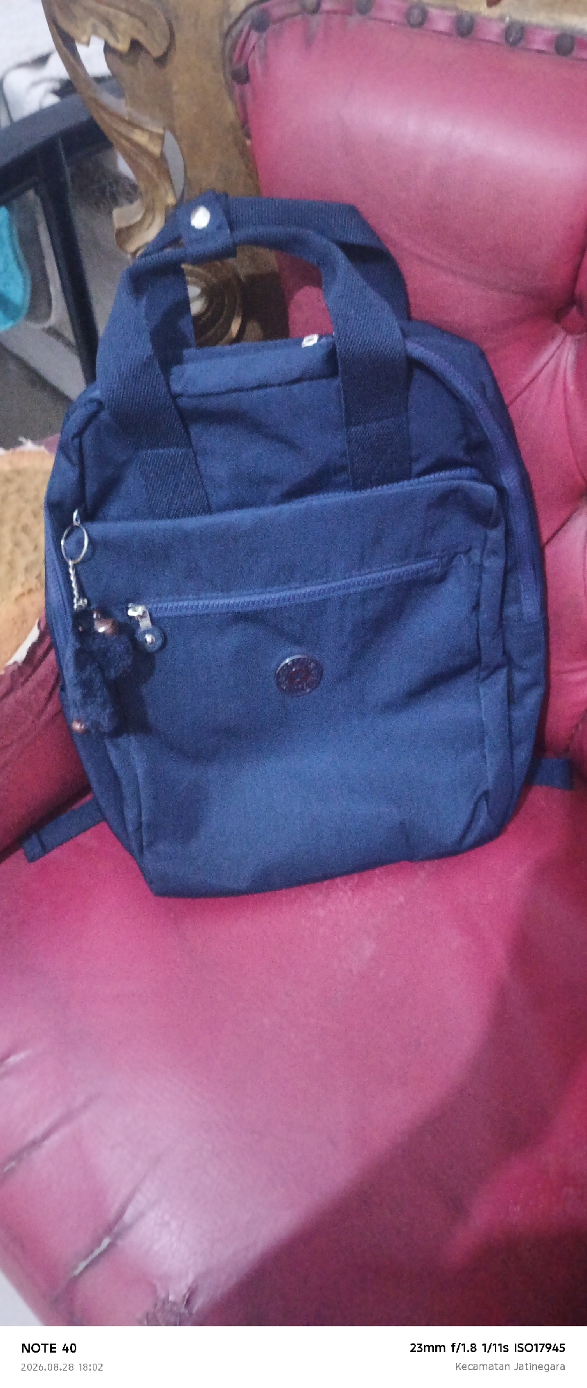 A dark blue backpack is centrally placed on a plush, magenta-colored chair. The backpack has a front zippered pocket and a circular emblem near the bottom of the front panel. A keychain with a fuzzy brown charm hangs from the zipper pull of the front pocket. The chair appears to be made of leather or a similar material and shows signs of wear, with some creases and scuffs visible. In the background, a portion of an ornate wooden furniture piece is visible on the left side of the image. The bottom of the image contains text overlay with information including "NOTE 40", a date and time "2026.08.28 18:02", and camera settings "23mm f/1.8 1/11s ISO17945" followed by "Kecamatan Jatinegara".