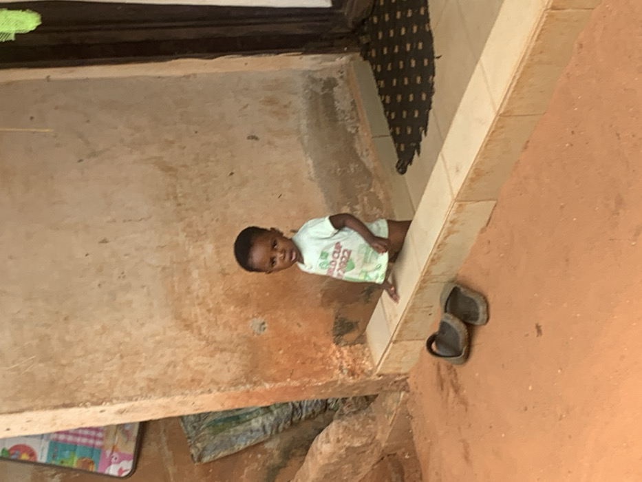 The scene is set in Otuocha, Nigeria. A young child, appearing to be a toddler, stands looking directly forward from within an entrance or recessed area. The child has dark skin and short dark hair, and wears a light green short-sleeved t-shirt featuring "WORLD CUP 2022" text and a graphic. The child's left hand rests on a low, light-colored tiled ledge that forms the threshold of the entrance. In the foreground, on a reddish-brown dirt ground, two dark-colored open-toe sandals are positioned to the left of the ledge. The background consists of a textured, light brown wall exhibiting signs of wear and discoloration. To the far left, a colorful patterned mat hangs partially visible on the wall, with another folded item below it. To the right, a dark wooden doorframe is visible, with a dark, patterned doormat placed on the floor inside the threshold. The area is illuminated by natural light.