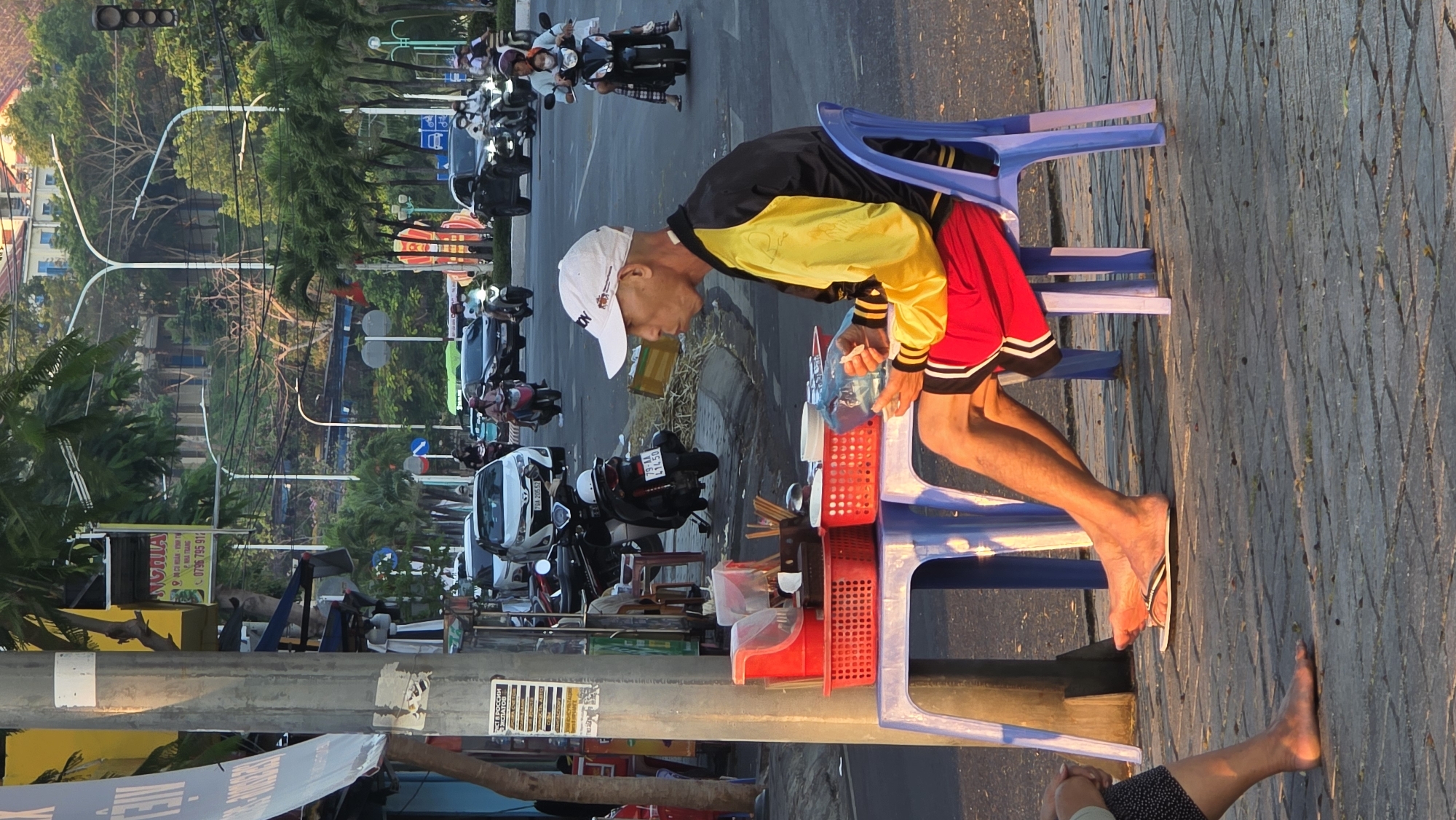 A vendor sits on a blue plastic stool at a makeshift street food stall on a brick sidewalk in Bắc Nha Trang, Vietnam. The vendor, wearing a white baseball cap, a black and yellow jacket, and red shorts, prepares food. The stall consists of plastic baskets and containers holding utensils and ingredients. In the background, a busy street is filled with motorbikes and cars. Trees line the street. Streetlights are visible, as are signs in Vietnamese. Another person's foot is seen in the lower right corner of the image.