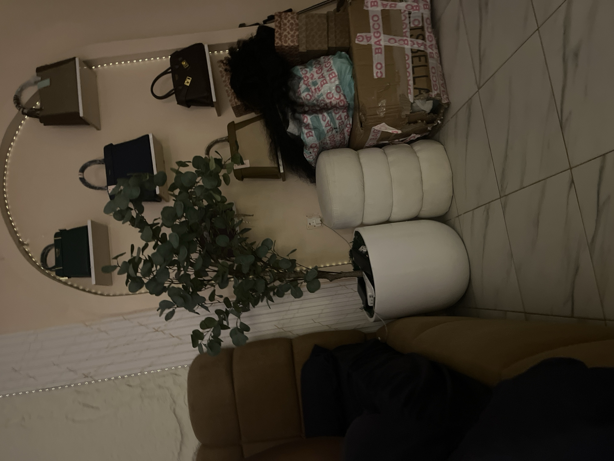 This is a medium-shot, eye-level, indoor photograph of a room with a collection of handbags displayed on shelves. The room appears to be a domestic setting in Abuja, Nigeria, likely a bedroom or dressing area, judging by the presence of a plush sofa and the overall decor. The lighting is dim, suggesting it is evening or nighttime, with artificial light sources casting soft shadows.
The focal point of the image is a wall with a curved archway, illuminated by a string of small lights along its perimeter. Mounted on shelves within this archway are several handbags of varying colors and sizes. From top to bottom and left to right, the visible bags include a dark green structured handbag on the top left shelf, a taupe-colored handbag with light blue accents on the top right shelf, a navy blue structured handbag on the middle left shelf, and a dark brown structured handbag with gold hardware on the middle right shelf.
In the foreground, to the left, a portion of a large, brown, tufted sofa is visible, with a dark blue cushion or blanket draped over it. To the right of the sofa and partially obscuring the lowest shelf is a tall, white, cylindrical planter containing a large artificial plant with numerous green leaves.
In front of the planter and to the right, a stack of four round, white poufs is visible. To the right of the poufs, there are several cardboard boxes of various sizes, some stacked and some in disarray. One of the boxes is partially open and appears to contain items wrapped in plastic bags with the repeated text "BAGCO" printed on them in turquoise and pink. Another box on the far right is stacked with smaller, patterned boxes. There is also a fluffy black object, possibly a wig or faux fur throw, partially visible on top of the boxes.
The floor is tiled with large, light-colored tiles that have a marble-like pattern. The overall impression is one of organized clutter, with a focus on fashion accessories and a comfortable, lived-in atmosphere. No people are visible in the image.