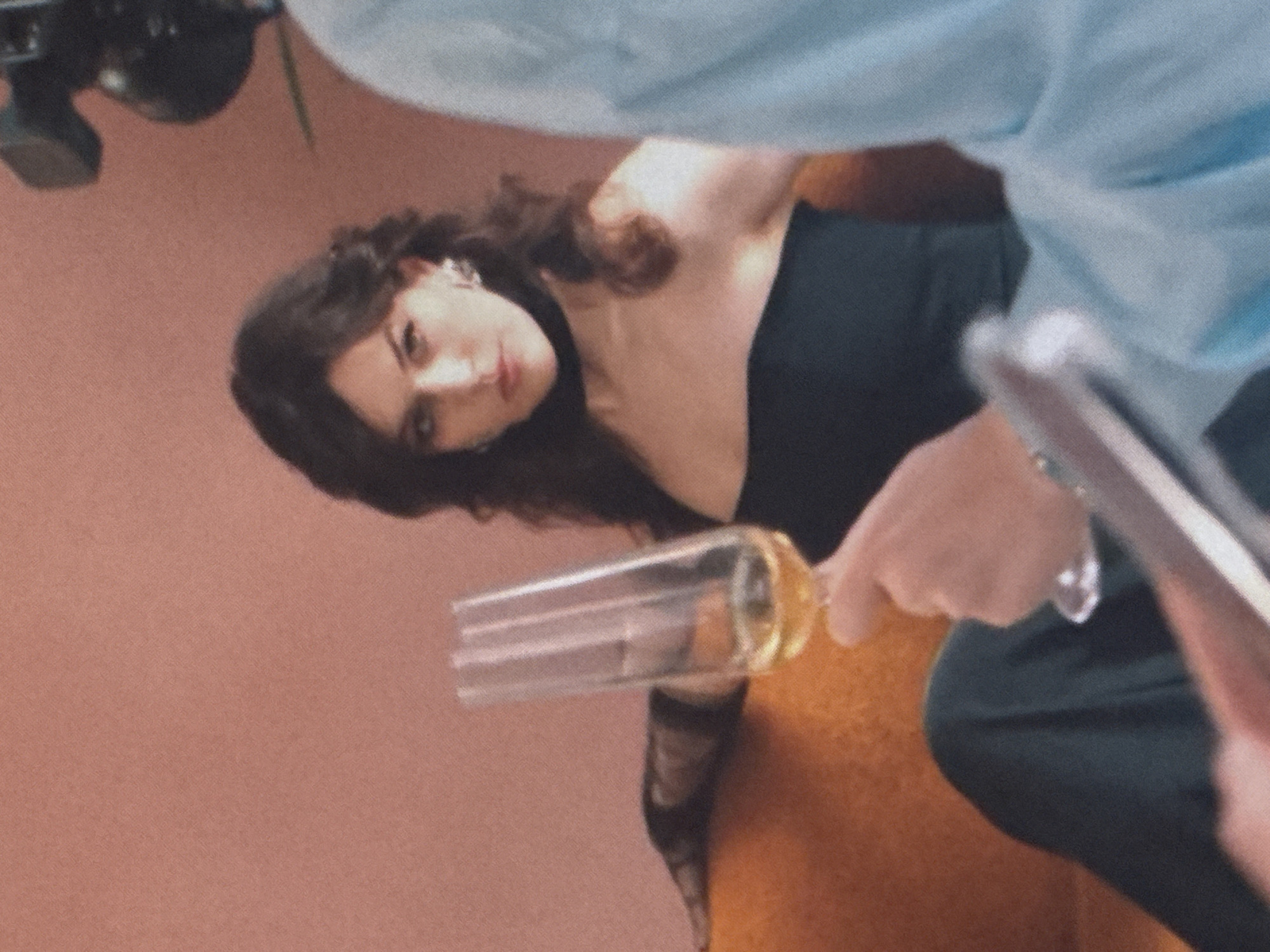 A woman with dark hair and prominent earrings is seated on an orange couch, holding a champagne flute filled with a pale yellow liquid. She wears a black off-the-shoulder top and a black choker necklace. Her gaze is directed towards the viewer. To the right of the woman, a person wearing a light blue garment is visible, holding a camera, suggesting a photoshoot or filming. The background is a textured, peachy-pink wall. A blurred, dark object with a striped pattern rests on the couch behind the woman. The overall lighting is soft and diffused. This scene is not associated with Saratov, Russia.
