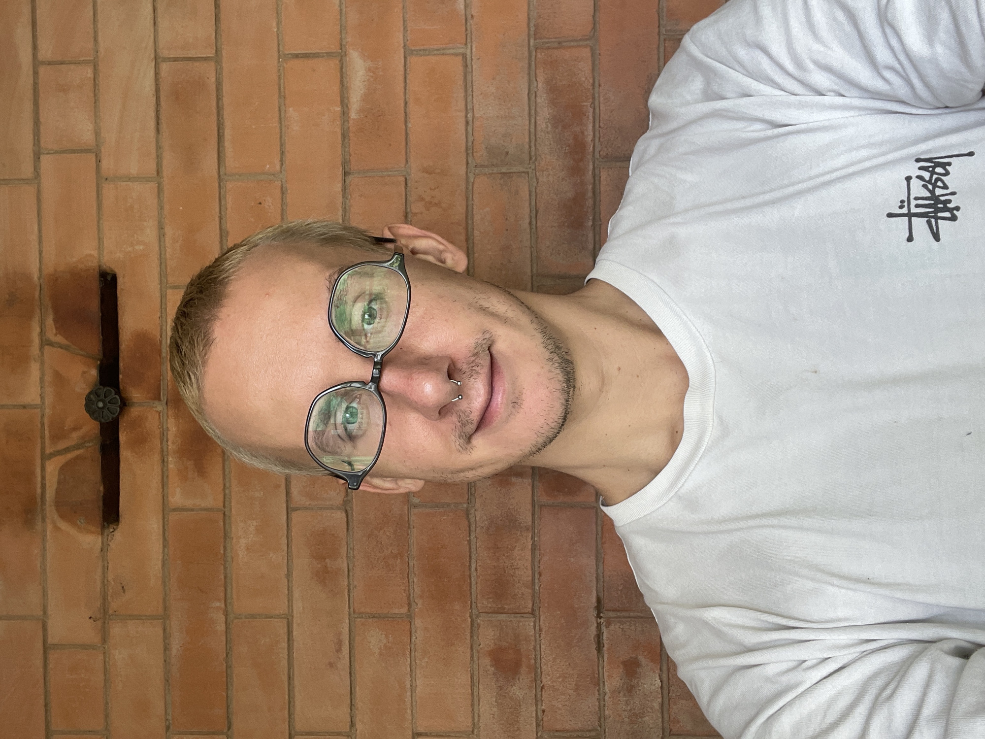 A fair-skinned male with short, blonde hair and a neatly trimmed mustache and stubble is posing in front of a red brick wall. He is wearing round, black-framed glasses and a white long-sleeved shirt with a black logo. He has a piercing in his nose. A dark, vertical object with a decorative circular element is affixed to the brick wall to his left. The photo appears to be taken indoors. There is no evidence in the photo to indicate that this photo was taken in Sestroretsk, Russia.