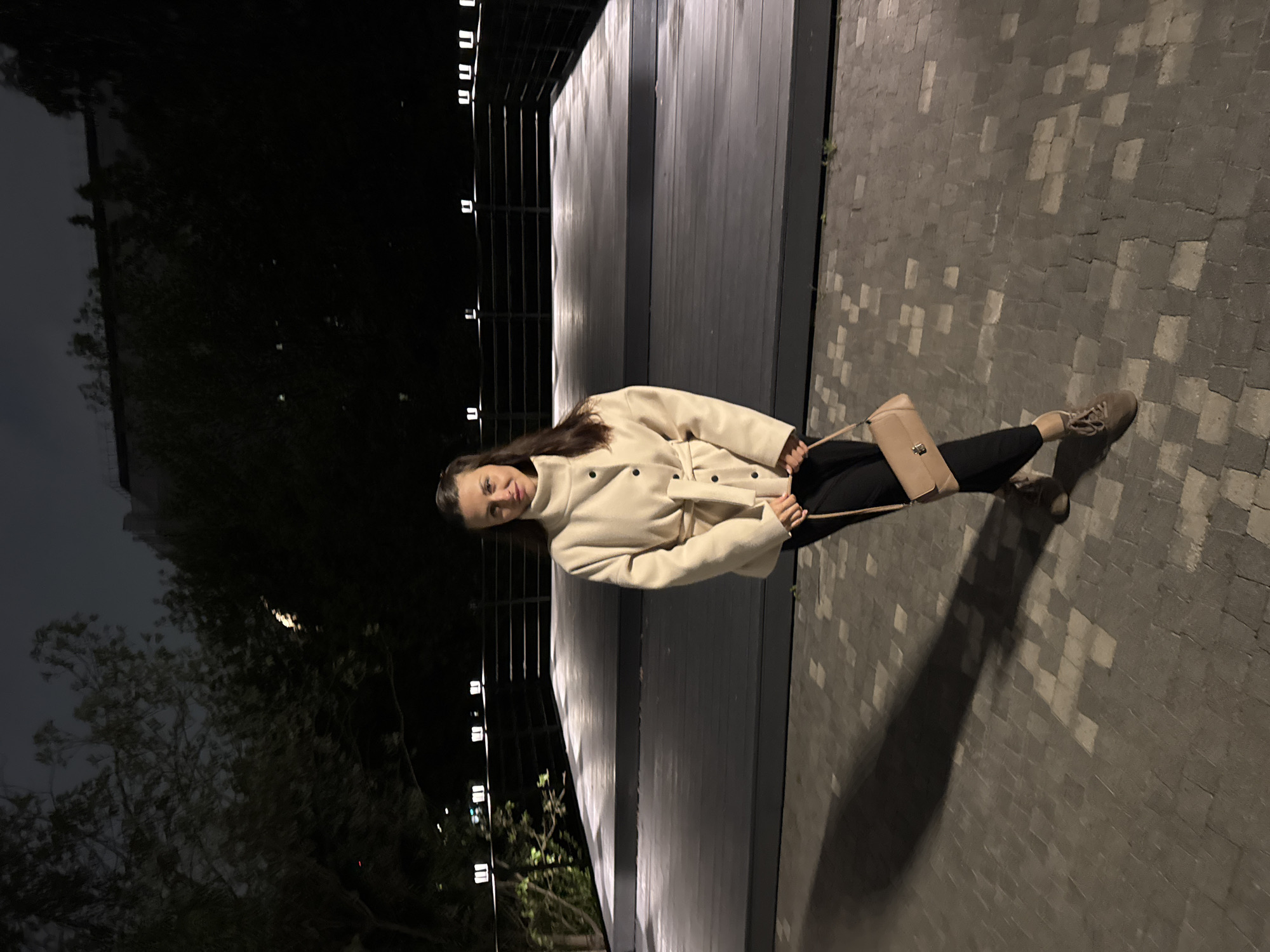 A woman stands at night on a sidewalk made of gray cobblestones, with a wooden structure featuring a railing to her left. The structure is illuminated by small, square lights. Behind the structure, trees and a building are visible in the darkness. The woman is wearing a light-colored jacket, black pants, and light-colored shoes. She is holding a light-colored purse. She is looking towards the camera. The image is taken at a slight angle. Based on the style of the architecture, lighting, and the cobblestone sidewalk, the location is likely in Saint Petersburg, Russia.