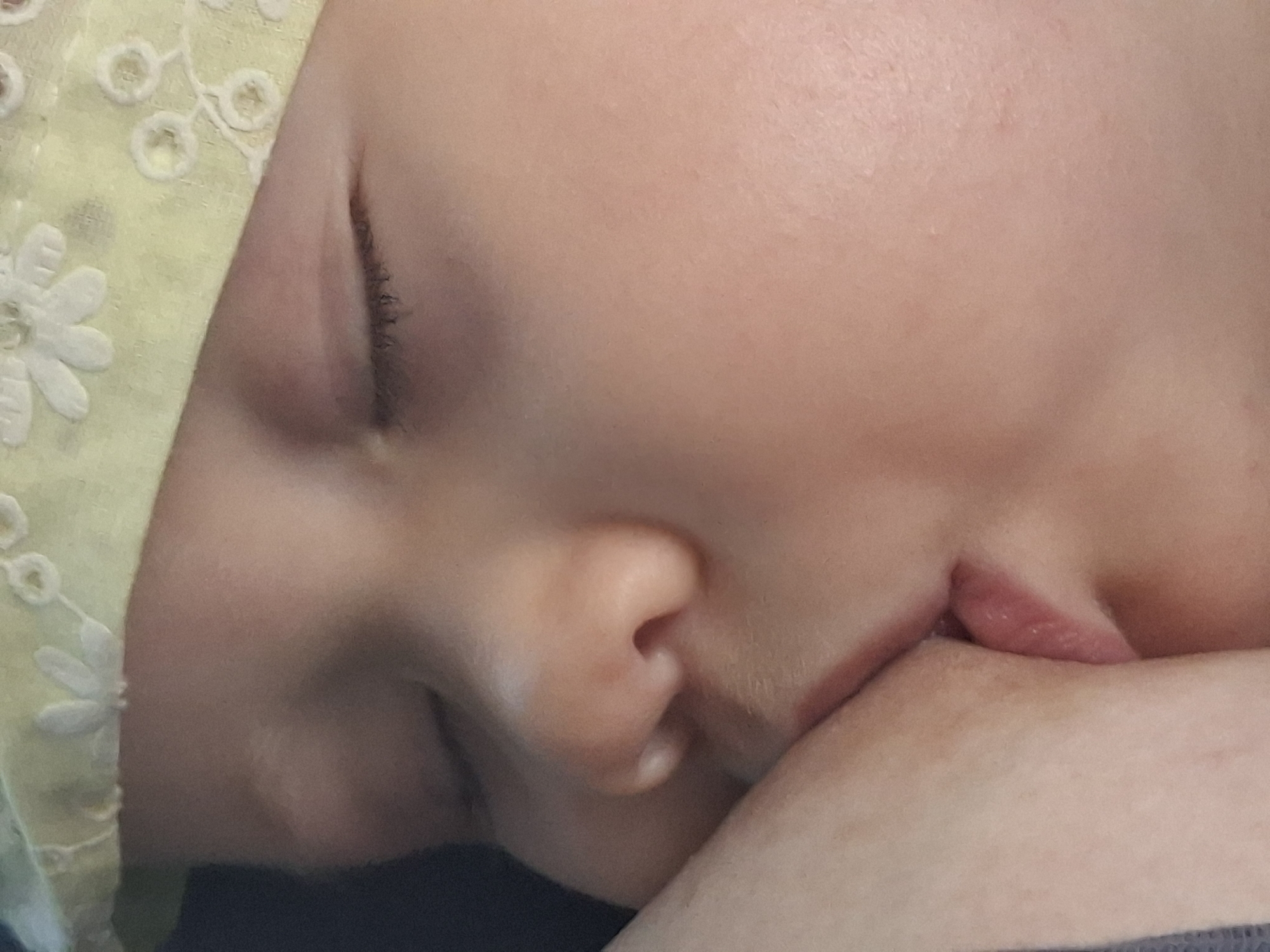 The image is a close-up, overhead shot of a baby's face and chest, with a portion of an adult's arm and breast visible. The baby is wearing a light yellow bandana with white floral embroidery. The baby's eyes are closed, and their mouth is latched onto the adult's breast. The background is dark and out of focus. There is no indication of the city of Semey, Kazakhstan, or any urban environment in the image.