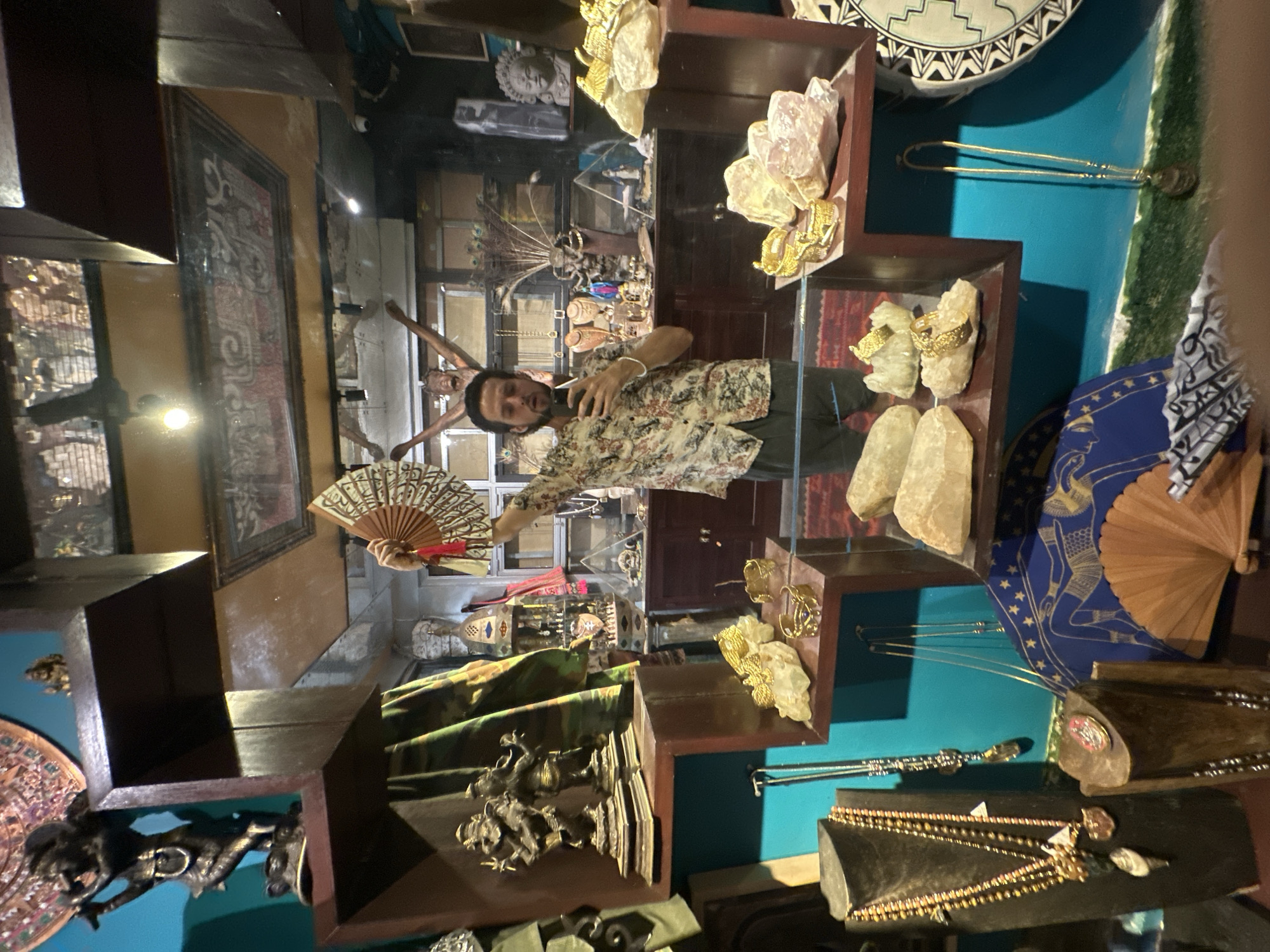 A man is standing in a shop filled with jewelry and statues, holding up a fan and taking a selfie. The shop is decorated with various religious and cultural artifacts, including statues of Hindu deities and Buddhist figures. There are also shelves filled with necklaces, bracelets, and other decorative items. The man is wearing a floral shirt and dark shorts. He is in the center of the image, with the shop's reflection visible in the mirror behind him. The shop appears to be located in Ubud, Indonesia, judging by the religious artifacts and the overall ambiance.