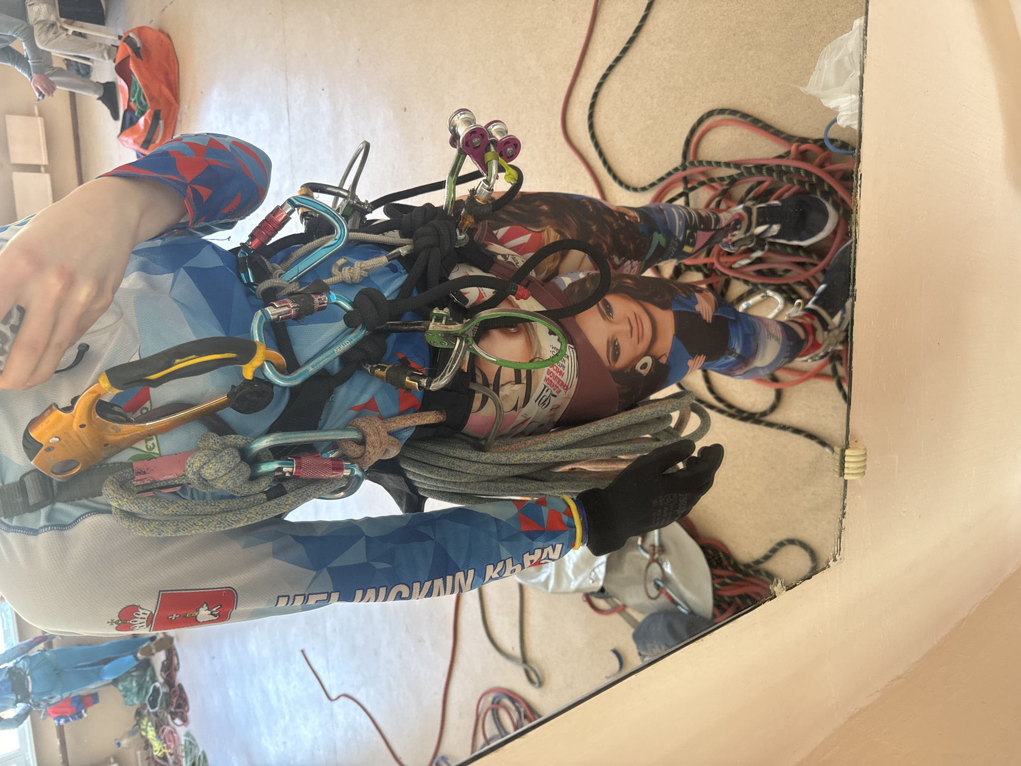 A high-angle, indoor shot shows a person outfitted in climbing gear, positioned in front of a large mirror reflecting the scene. The individual wears a blue and white long-sleeved shirt adorned with red geometric patterns and the Polish coat of arms. Their climbing harness is laden with an array of carabiners, ropes, and other climbing equipment, including a yellow-handled rigging plate.
Strangely, the harness also supports a rolled-up magazine or poster featuring an image of a woman with dark hair. The floor is covered with loose coils of red and black ropes, and a pair of black sneakers are visible among them.
The reflection in the mirror reveals additional details: A second person is partially visible in the background, along with an orange backpack and some wooden furniture. The walls are painted in a pale neutral color. The location is likely a training facility for climbing or a related sport, potentially in Usolye, Russia, given the Polish coat of arms and clothing text which may be Russian.