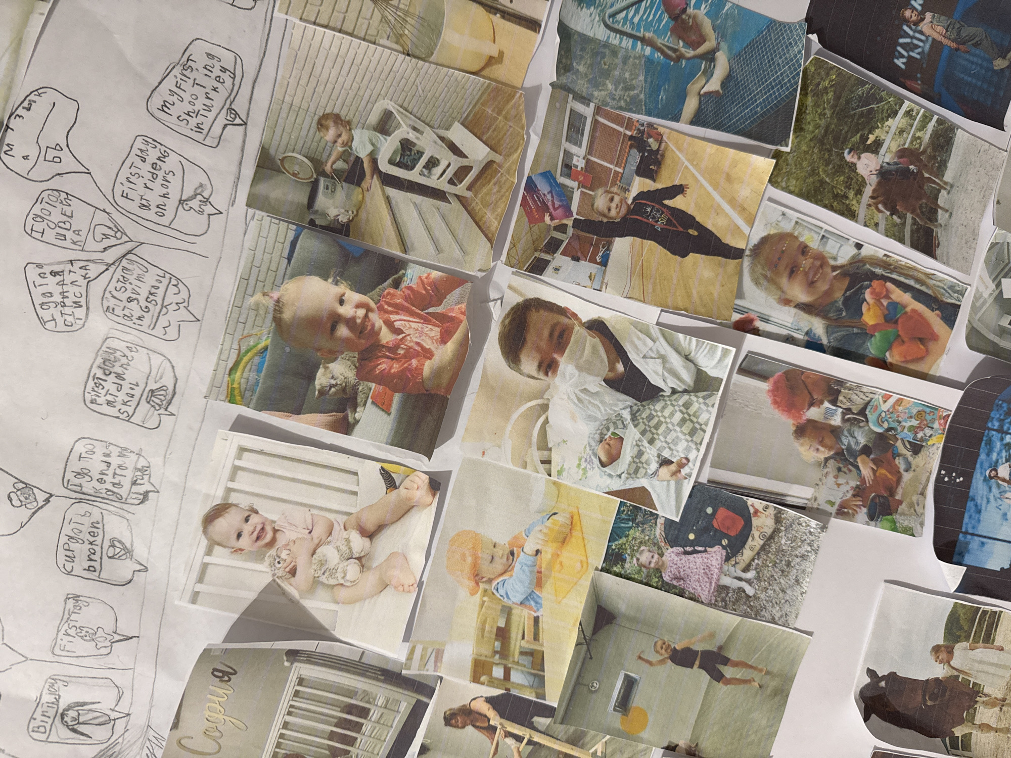The image shows a collection of printed photographs and a hand-drawn sheet of paper arranged on a white surface, possibly a table. The photographs depict a variety of subjects, mostly appearing to be of a young girl at different stages of her life, including baby pictures in a crib, playing, swimming, riding a horse, and dressed in a graduation gown.
The hand-drawn sheet includes speech bubbles with text in what appears to be Russian. The text includes phrases such as "Birthday," "First Toy," "Cup gets broken," "I do too," "First day at school," "I go to," "First day riding on horse," and "My first shooting in Turkey." The layout suggests an attempt to narrate or caption the photographs.
The overall scene gives the impression of a personal photo album or scrapbook in the process of being created or reviewed. The arrangement is somewhat scattered, suggesting an informal and personal setting. The environment appears to be indoors, with the focus entirely on the photographs and drawing. There is no indication of the city of Budva, Montenegro, in the image.