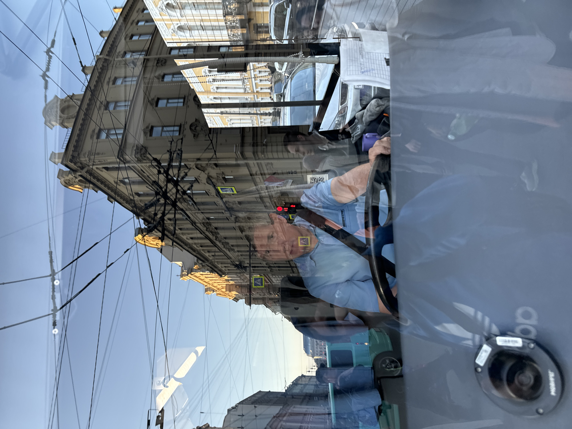 The image is a reflection of a street scene in Saint Petersburg, Russia, seen through the windshield of a vehicle. The driver, a man wearing a light blue shirt and a black seatbelt, is visible behind the steering wheel. The reflection shows classic Saint Petersburg architecture with ornate building facades, street signs, and overhead trolley wires. In the foreground, part of the vehicle's interior is visible, including a horn and some papers. The overall impression is that of a sunny day in the city, with the reflections creating a layered and complex visual.