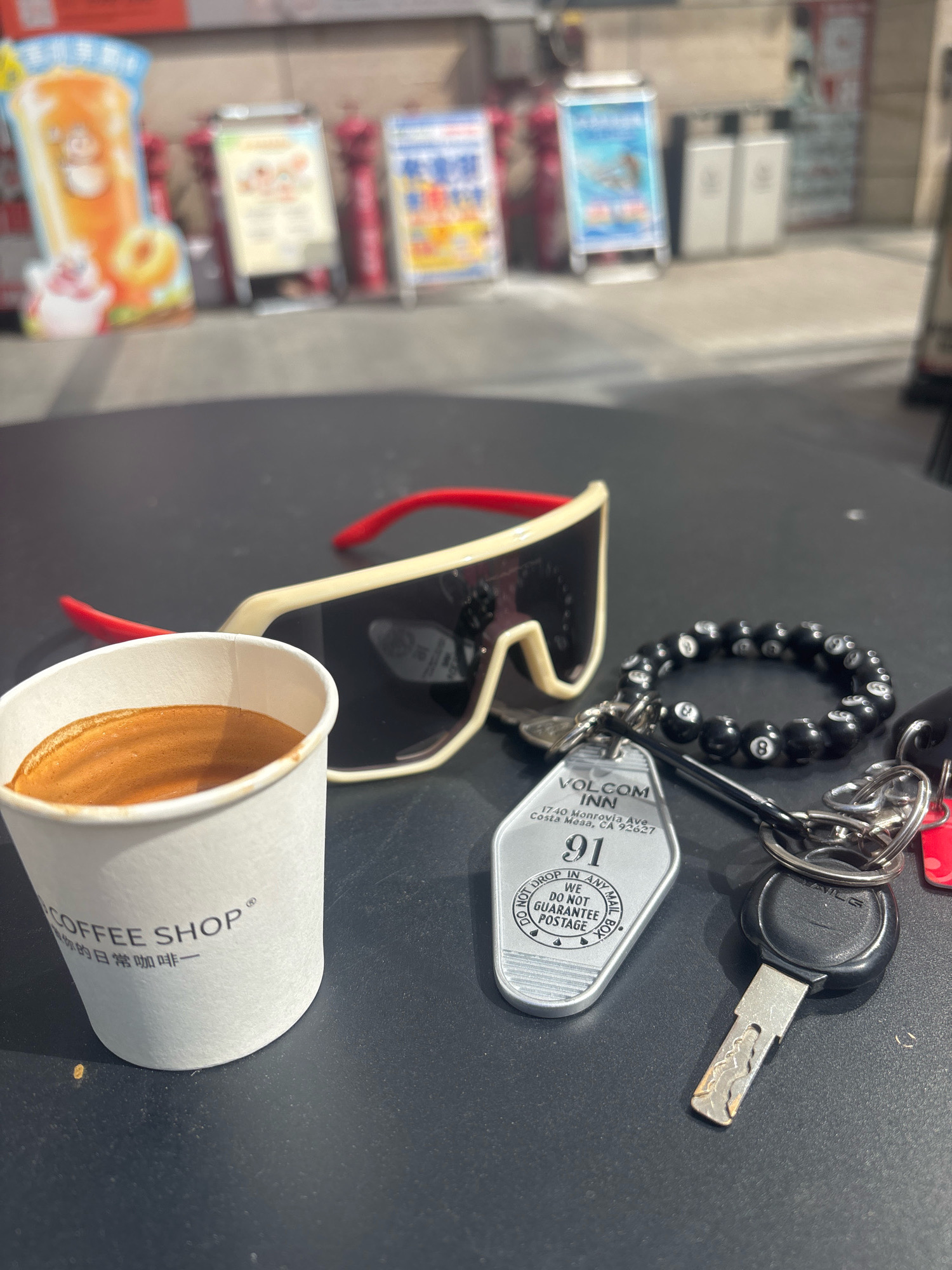 On a dark, reflective surface, possibly a table at an outdoor cafe, several objects are arranged. A white paper cup, partially filled with a dark liquid topped with foam, likely espresso, sits on the left. The cup has text printed on it, including "COFFEE SHOP" and Chinese characters. To the right of the cup are a pair of large, white and red-rimmed sunglasses with dark lenses. Attached to a keyring with multiple keys is a silver hotel room key tag for "VOLCOM INN" located at "1740 Monrovia Ave, Costa Mesa, CA 92627," with the room number "91" prominently displayed. A black beaded bracelet, adorned with what appear to be billiard ball motifs, is also part of the keyring assembly. The background is blurred but suggests an outdoor setting with colorful advertisements and possibly a street or walkway. There are no people visible in the image. The lighting suggests it is daytime. The overall scene captures a collection of personal items in a casual, urban environment. The mention of Nanhai District, China in the prompt is not supported by any visual evidence within the image itself.