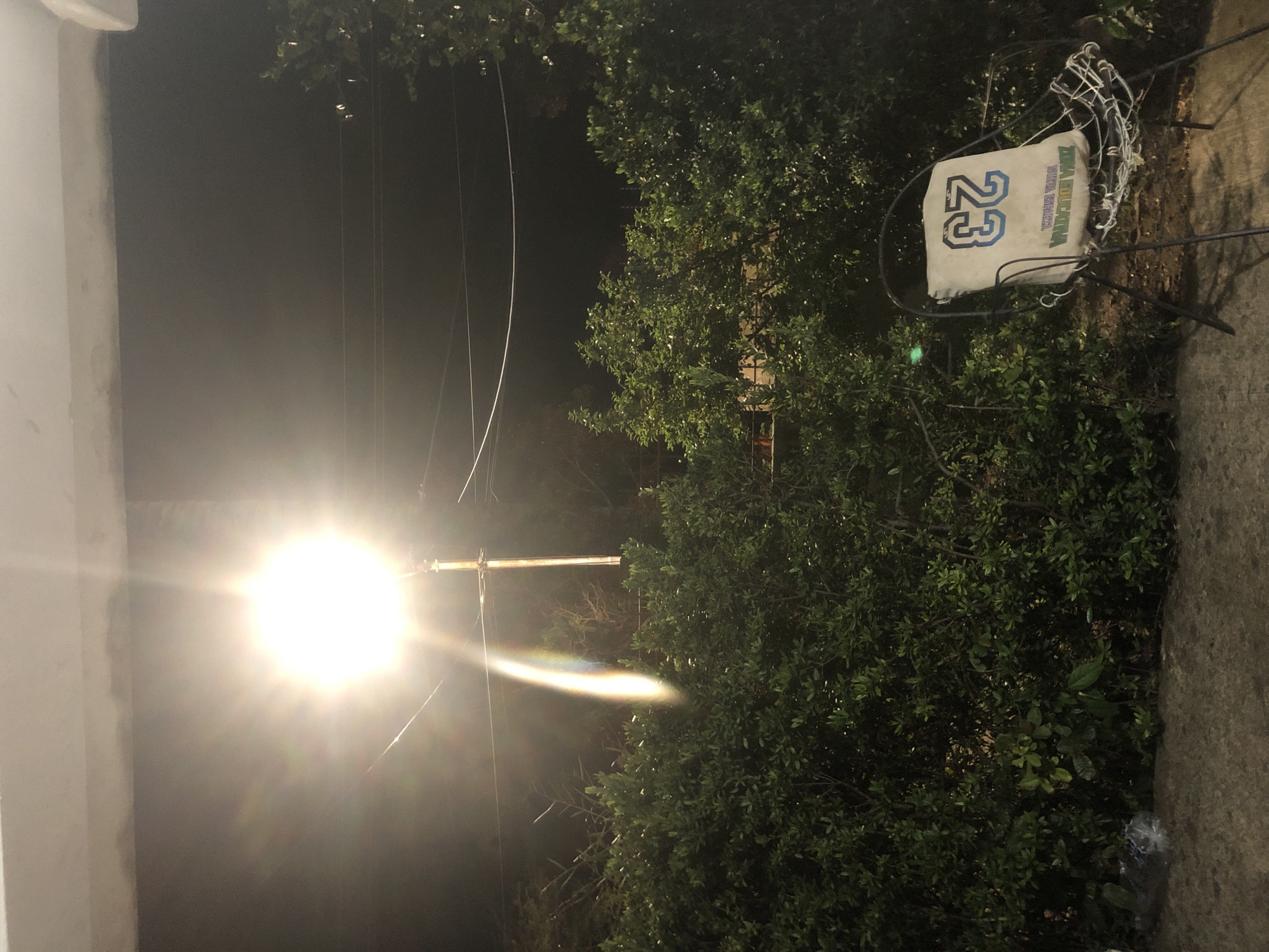 A wide shot captures an outdoor scene at night. A bright overhead light illuminates the upper portion of the image, casting a rainbow-like sheen onto a lamppost behind it. Below the light, a dense hedge of dark green bushes stretches across the frame. To the right, a wire-frame chair sits on a concrete surface, with a pillow resting on it. The pillow is light-colored with the number "23" emblazoned on it in blue and light blue. There are no people visible in the frame. The environment appears to be a residential or public outdoor space.