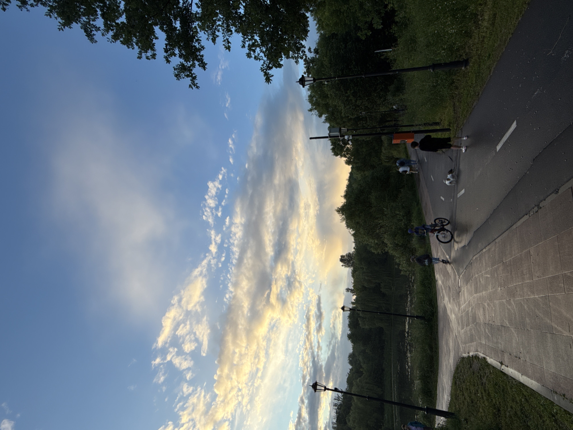 The image depicts a park scene in Moscow, Russia, likely during the late afternoon or early evening, given the soft, warm lighting and the presence of scattered clouds tinged with yellow and orange. The sky is a vibrant blue, with dynamic cloud formations suggesting a transition in weather.
In the foreground, a paved pathway curves through the park. To the left, the path is made of large square tiles, bordered by a grassy area. To the right, the path transitions to asphalt and appears to be a designated cycling or walking lane, marked with white lines.
Several individuals are present in the scene, engaged in recreational activities. A child on a bicycle is riding on the asphalt path. Further along the path, a young person is walking a dog. In the distance, a few other figures can be seen walking, suggesting a public park environment.
The park is lush with greenery, with a dense line of trees bordering a body of water, possibly a lake or pond, visible in the mid-ground. Several lampposts are strategically placed along the pathways, indicating the park is maintained for public use, including potentially after dark. The overall atmosphere is serene and suggests a pleasant outdoor setting.