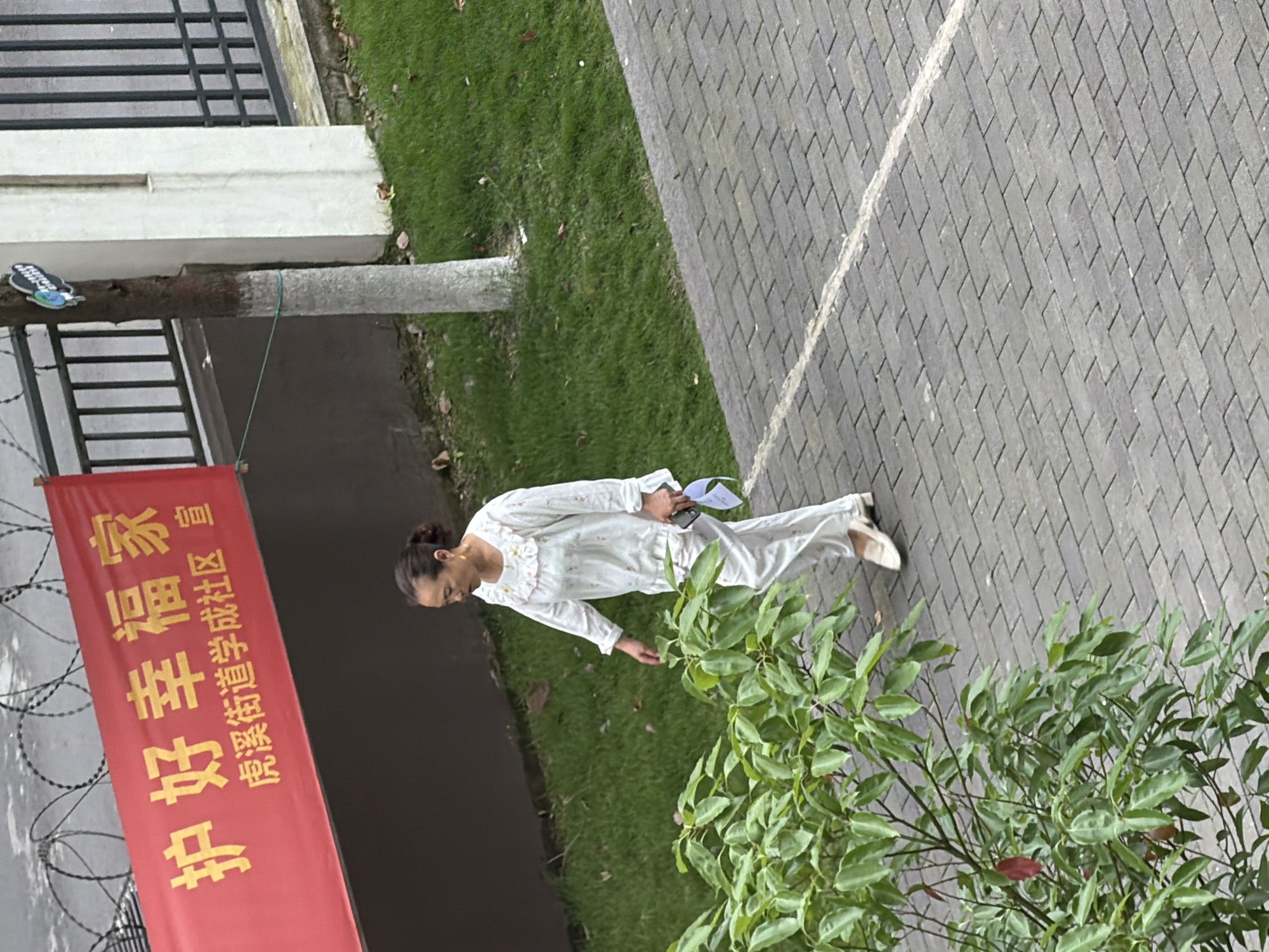 A woman wearing white pajamas stands near a bush on a paved area next to a grassy area in what appears to be an urban environment. A red banner with gold Chinese characters is visible on a pole, and the text " Chongqing High-tech Zone" is partially visible. A building with a metal fence and barbed wire is in the background. The woman is holding a cell phone and paper.