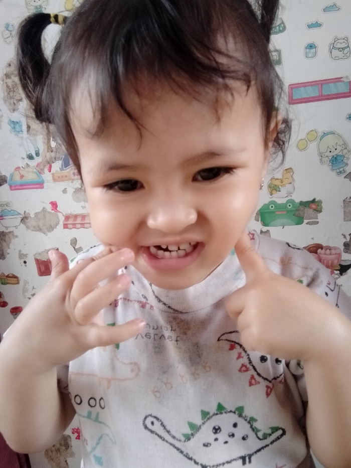 A toddler with dark hair in pigtails smiles, showing teeth. They wear a white t-shirt with dinosaur print. The child's right hand touches their cheek, while the left hand is held up near their face. The background is a wall covered in cartoon stickers and a peeling, textured pattern. The overall scene has a cheerful, playful vibe.