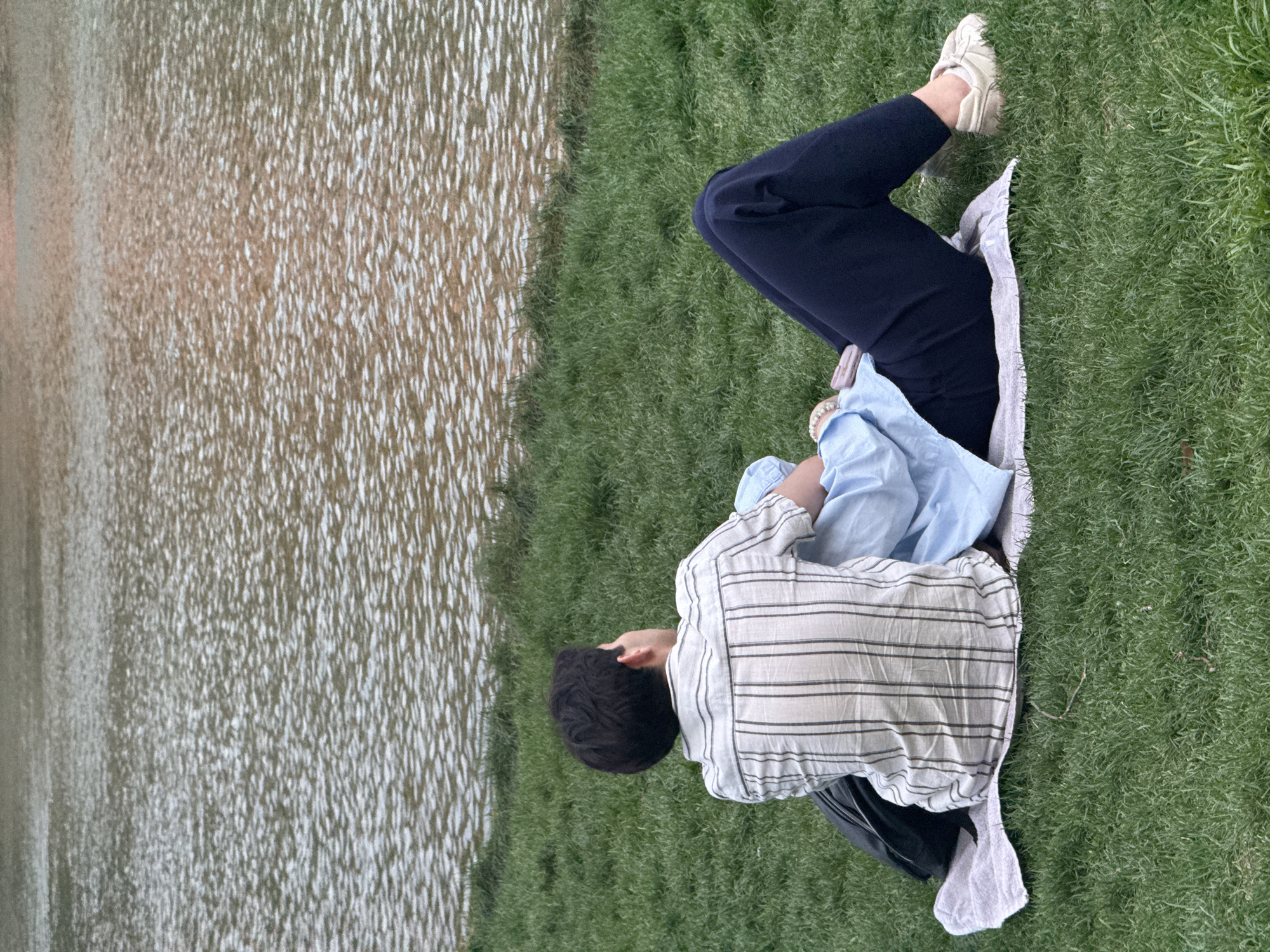 A person lies on a patch of green grass next to a body of water. The person is lying on their side, with their legs bent at the knees. They are wearing dark-colored pants and off-white shoes. They are also wearing a horizontally striped shirt and a light blue shirt. The body of water appears to be rippled. The location is unknown.