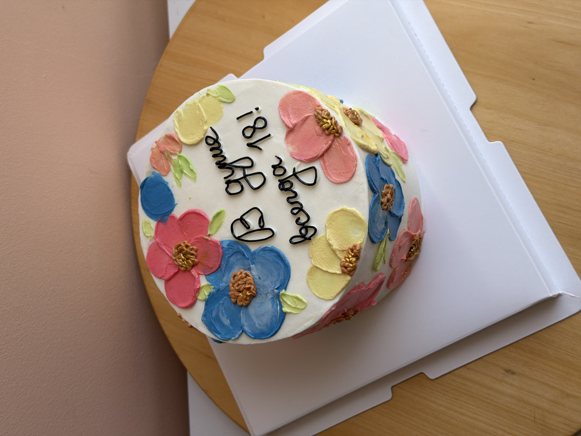 The image shows a round birthday cake in a white cardboard box, sitting on a wooden surface. The cake is decorated with white frosting, colorful buttercream flowers (pink, blue, yellow) and green leaves around the edges. There is black lettering on top of the cake saying “Happy Birthday” and “18!”. The backdrop includes part of a light pink wall and a light brown wooden surface. The location is not specified, so I cannot provide details about its situation in Tel-Aviv, Israel.