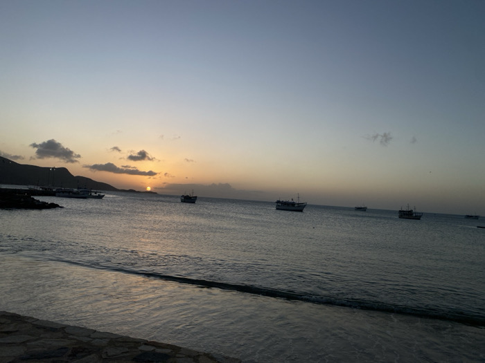 The image depicts a serene seascape at sunset. Several boats are anchored in the calm water. A low mountain range forms the backdrop under a sky transitioning from light blue to shades of orange and gray around the setting sun. The foreground shows gentle waves lapping against a stone barrier. The overall scene evokes tranquility and a quiet coastal atmosphere.