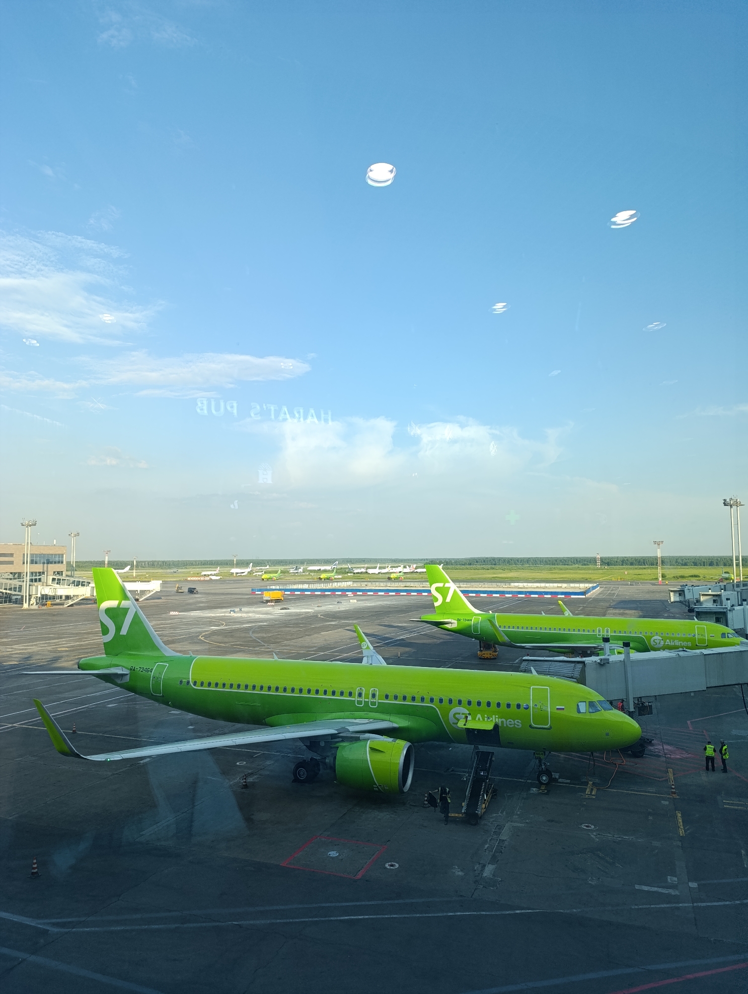 This media file captures an outdoor scene at an airport, likely Domodedovo, Russia, given the context. The main subjects are two bright green S7 Airlines airplanes parked on the tarmac. The foremost airplane, a passenger jet, is positioned closest to the viewer, its nose pointing towards the right. It is connected to a jet bridge, and a set of stairs is visible leading to the aircraft's door. Two ground crew members in high-visibility vests stand near the nose of this plane. Another S7 Airlines plane is parked behind it, with its tail visible.
The setting is an airport apron with a tarmac surface marked with various lines and symbols. In the background, other aircraft and airport infrastructure, including buildings and control towers, are visible. A line of trees forms the distant horizon under a bright, partly cloudy sky. The sun appears to be shining, suggesting it is daytime, possibly late morning or early afternoon, given the angle of the light and the presence of shadows.
Reflections are visible on what appears to be a window, distorting the view and adding an element of depth. Some of these reflections show abstract circular shapes, possibly from lights or other elements within the airport terminal. One reflection shows reversed text, which, when deciphered from the context, reads "PUBLICATION REPORT" in Cyrillic, indicating the viewing perspective is likely from inside a terminal building looking out.
The overall atmosphere is one of an active airport environment, with aircraft ready for or having completed flights. The vibrant green of the S7 Airlines livery stands out against the grey tarmac and blue sky.