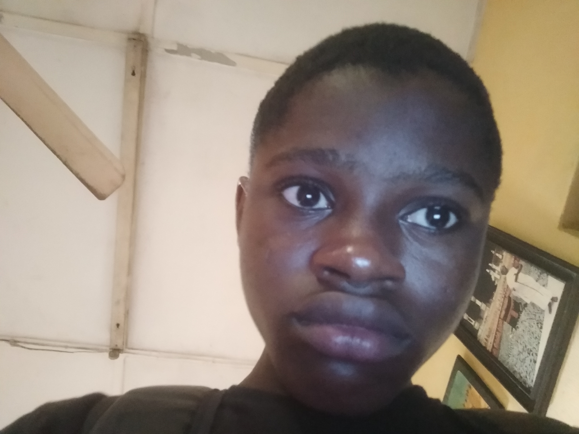 A close-up selfie shows a young person with dark skin and short, cropped hair. The person's face is centered in the frame, with a slight upward angle. The lighting is diffused, creating soft shadows. Behind the person, a white ceiling with visible wooden support beams can be seen, along with a ceiling fan. In the background, to the right, a yellow wall displays two framed pictures. The scene is indoors, possibly in a residential setting. The location in the city of Shangisha, Nigeria, is inferred.