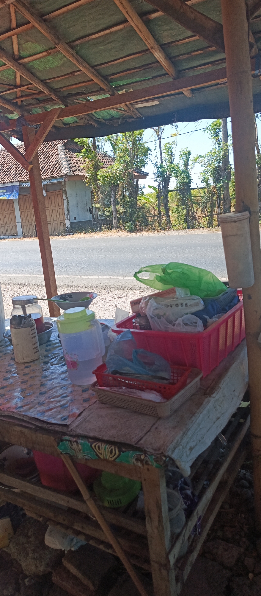 The image depicts an outdoor stall or makeshift shop located in Jatisari, Indonesia, beside a paved road. The stall is covered by a thatched roof supported by wooden beams, with some parts appearing weathered. In the foreground, a table is laden with various items. These include a patterned plastic tablecloth, a glass jar with a metal lid containing utensils, a bowl, a large plastic pitcher with a floral design filled with liquid, and several red and beige plastic baskets. The baskets contain an assortment of items, some wrapped in plastic bags, and one appears to hold fish. A green plastic bag is draped over the items in one of the baskets. In the background, a residential building with a tiled roof and closed wooden doors is visible, along with trees and vegetation lining the roadside. The overall scene suggests a simple, informal commercial setup.