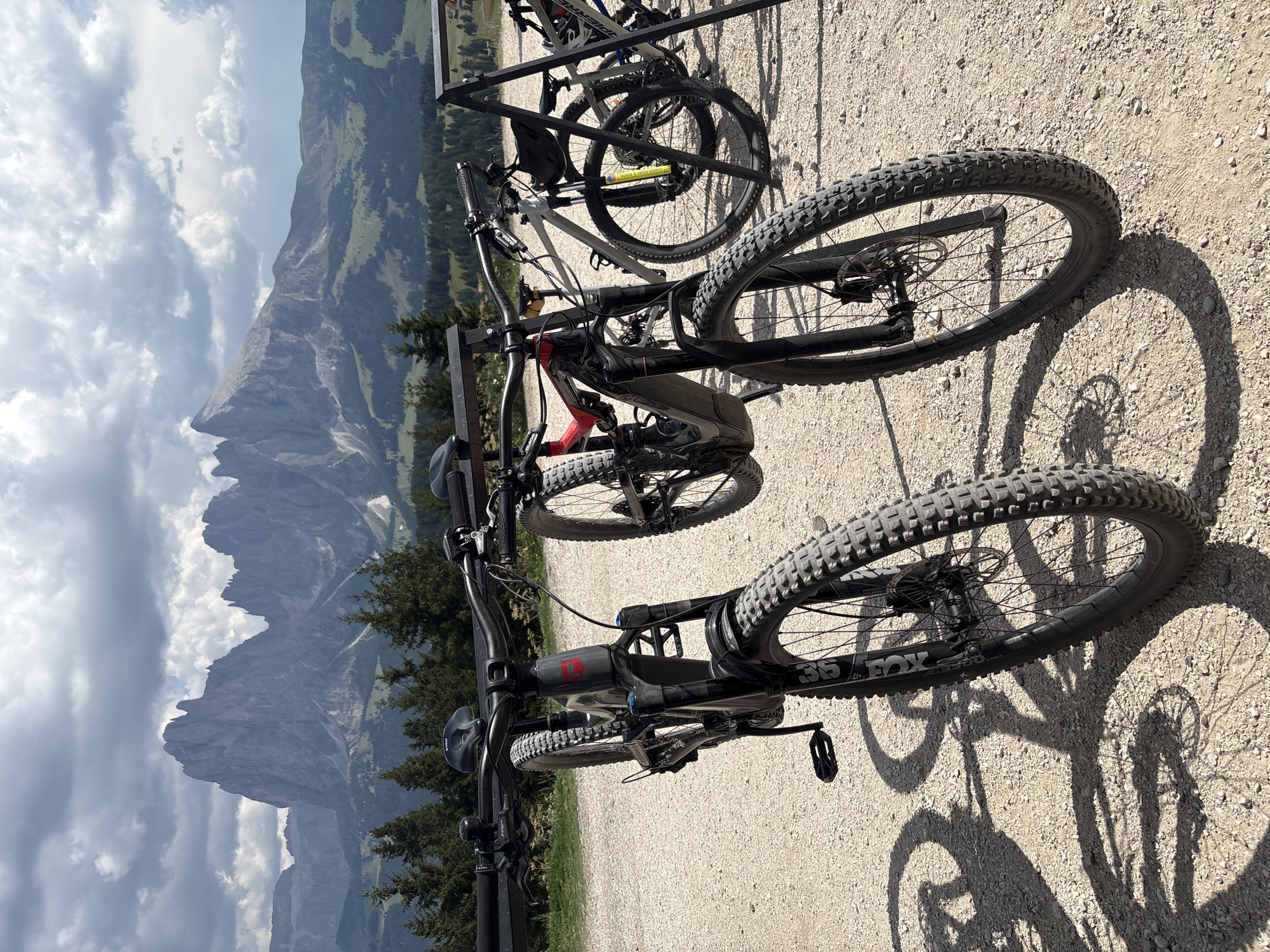 The image depicts a scenic mountainous landscape near Kastelruth - Castelrotto, Italy. In the foreground, two mountain bikes are parked on a gravel path. The bike on the left is black with red accents and has a prominent "5" logo on its frame. Its front wheel is angled, and its shadow stretches across the ground. The bike on the right is partially obscured but appears to be lighter in color. Behind the bikes, the landscape transitions from scattered trees to rolling green hills, culminating in the dramatic, jagged peaks of the Dolomites under a cloudy sky. The overall impression is one of outdoor adventure and natural beauty.