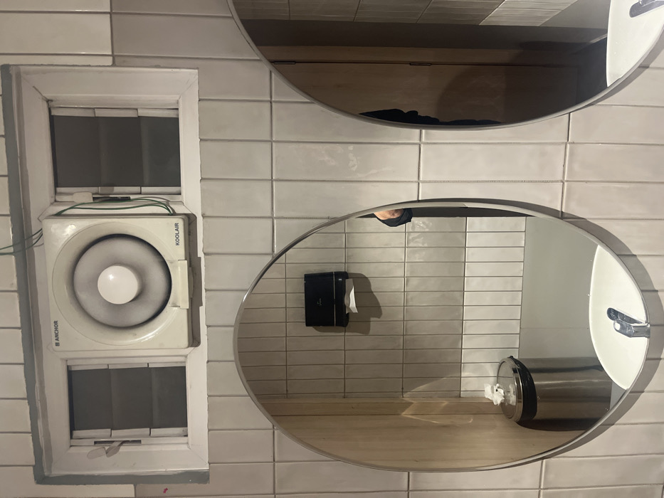 The image displays an interior scene, likely a public restroom or utility room in Dibrugarh, India, featuring light-colored, possibly white, subway-style tiled walls.
On the left side of the frame, there is a rectangular cut-out resembling a window. Within this opening, a white ventilation unit is installed, featuring a prominent circular fan. The unit is branded "KOOLAIR" on its upper right side and "ANCHOR" on its lower left. Green wiring is visible connected to the top of the unit. Below the fan, the lower part of the window opening reveals slatted blinds.
Mounted on the tiled wall to the right are two oval-shaped mirrors. The lower mirror reflects a black paper towel dispenser, a cylindrical silver bin (possibly a waste receptacle or another dispenser), a partial view of a white sink with a chrome faucet, and a wooden counter or floor. A person's foot, clad in a black slipper or sandal, is clearly visible in the reflection of this mirror. The upper mirror primarily reflects a wooden surface, which could be a door or another wall, along with some dark fabric and a faint reflection of the same person's foot. The presence of the foot in both mirrors indicates that the photographer is standing in front of them. The lighting suggests a well-lit indoor environment.