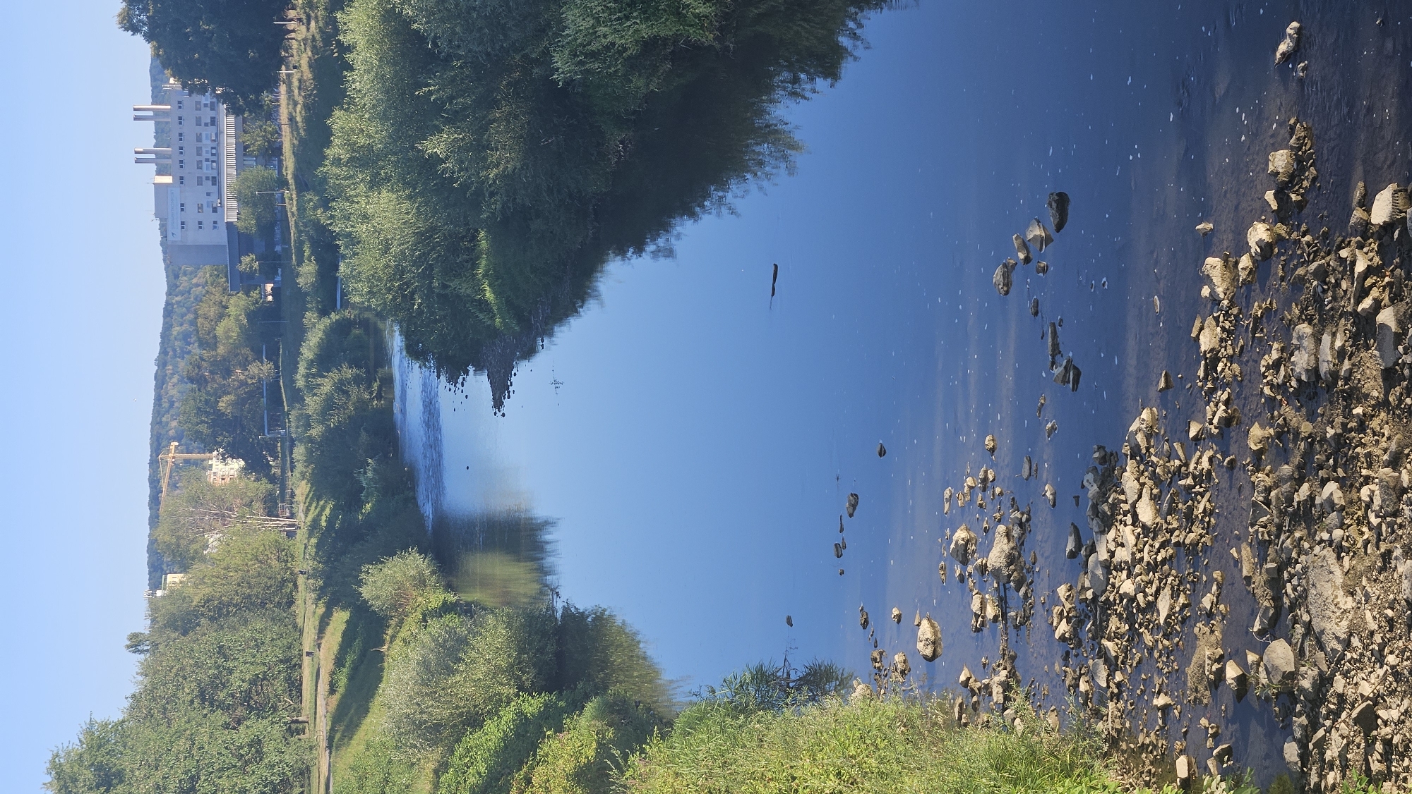 This image, taken in Prešov, Slovakia, depicts a calm river flowing through a vegetated area with buildings and hills in the background. The river is clear blue, reflecting the sky and surrounding trees. Rocks and pebbles are visible along the riverbed, particularly in the foreground. Lush green bushes and trees line the banks, casting shadows on the water. In the distance, a large, modern building with multiple chimneys stands out, along with a construction crane and rolling hills covered in trees. The sky is clear and bright blue, indicating a sunny day. There are no people or animals visible in the image.