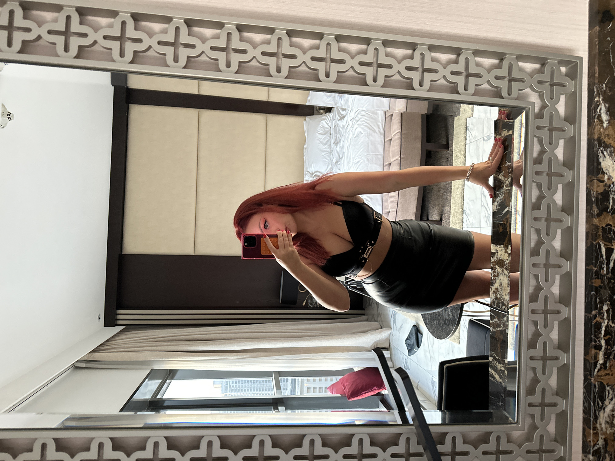 A woman with red hair is taking a mirror selfie in a hotel room. She is wearing a black leather crop top and a black leather mini skirt. She has on red lipstick and her nails are painted red. In the background, there is a large bed with white linens, a window with a view of a city skyline, and a mirror with a decorative frame. The room appears to be modern and luxurious. The city visible outside the window could be Dubai, United Arab Emirates, due to the style of the skyscrapers.