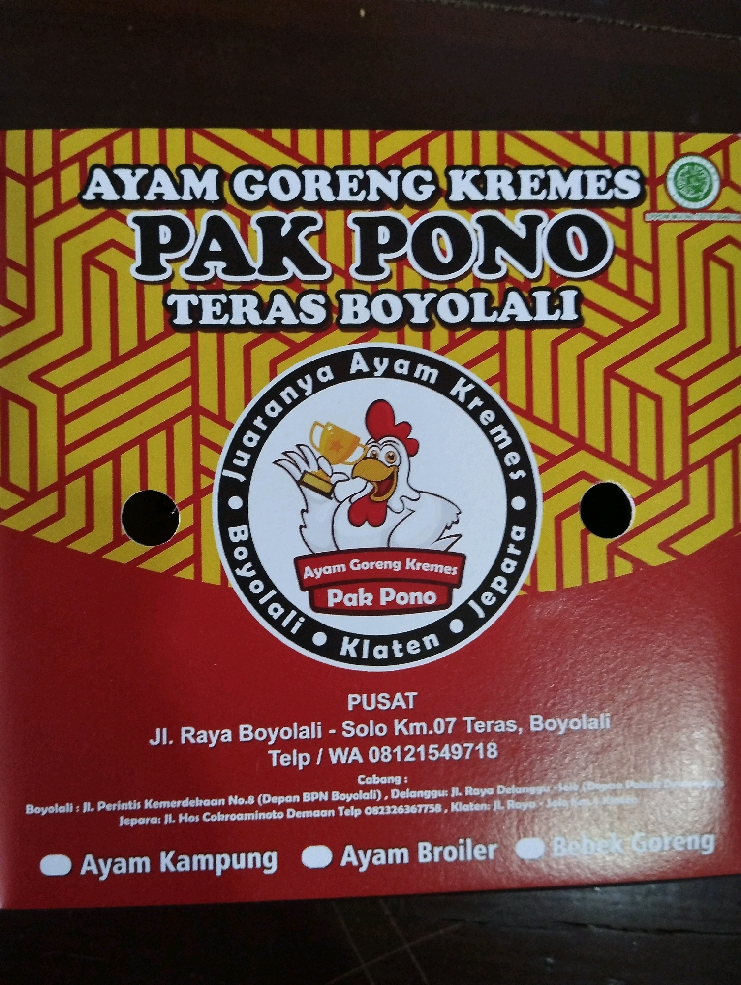 The image is a product label or packaging for "AYAM GORENG KREME" from "PAK PONO". The label features a cartoon chicken holding trophies, a circular design with text "JUARANYA Ayam Kremes Boyolali Klaten Jepara". Below the circular design, it lists the "PUSAT" (Center) location as "Jl. Raya Boyolali - Solo Km.07 Teras, Boyolali" with a phone number "Telp / WA 08121549718". It also provides "Cabang" (Branch) information for Boyolali and Jepara. At the bottom, it lists menu items: "Ayam Kampung", "Ayam Broiler", and "Bebek Goreng". The image is taken from a slightly overhead angle, and the overall color scheme is red, yellow, and white with black text. There are two black circular cutouts, possibly for hanging or display. The background appears to be a wooden surface. The product is not in Salatiga, Indonesia, but rather from Boyolali.