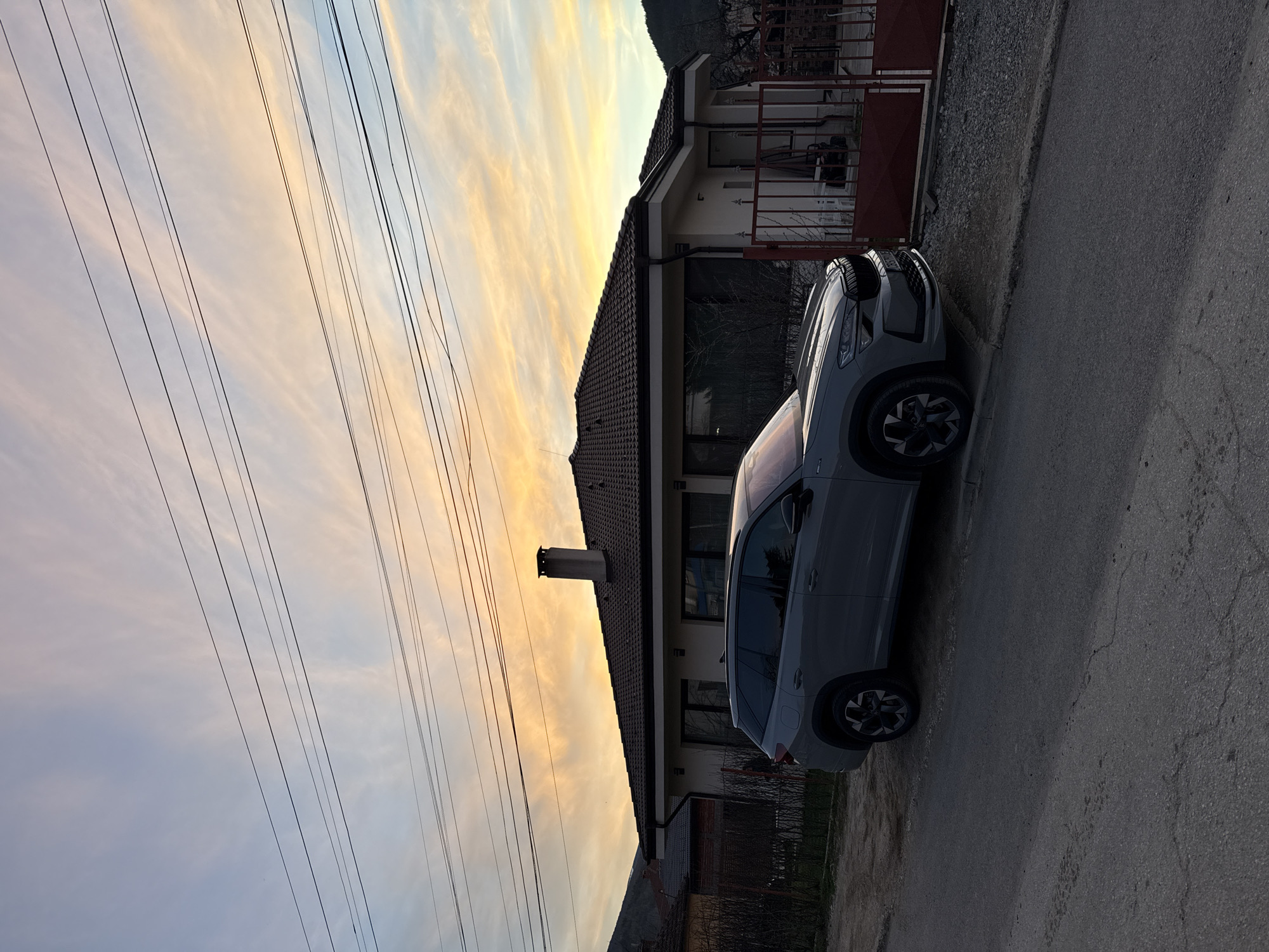 The image shows a gray car parked on the side of a road in Etropole, Bulgaria. A two-story house stands next to the car, with a gate in front. Overhead power lines stretch across the sky, which displays a mix of light orange, yellow, and blue hues, suggesting sunset or sunrise. The road appears to be asphalt. The scene is slightly angled, with the sky positioned vertically.