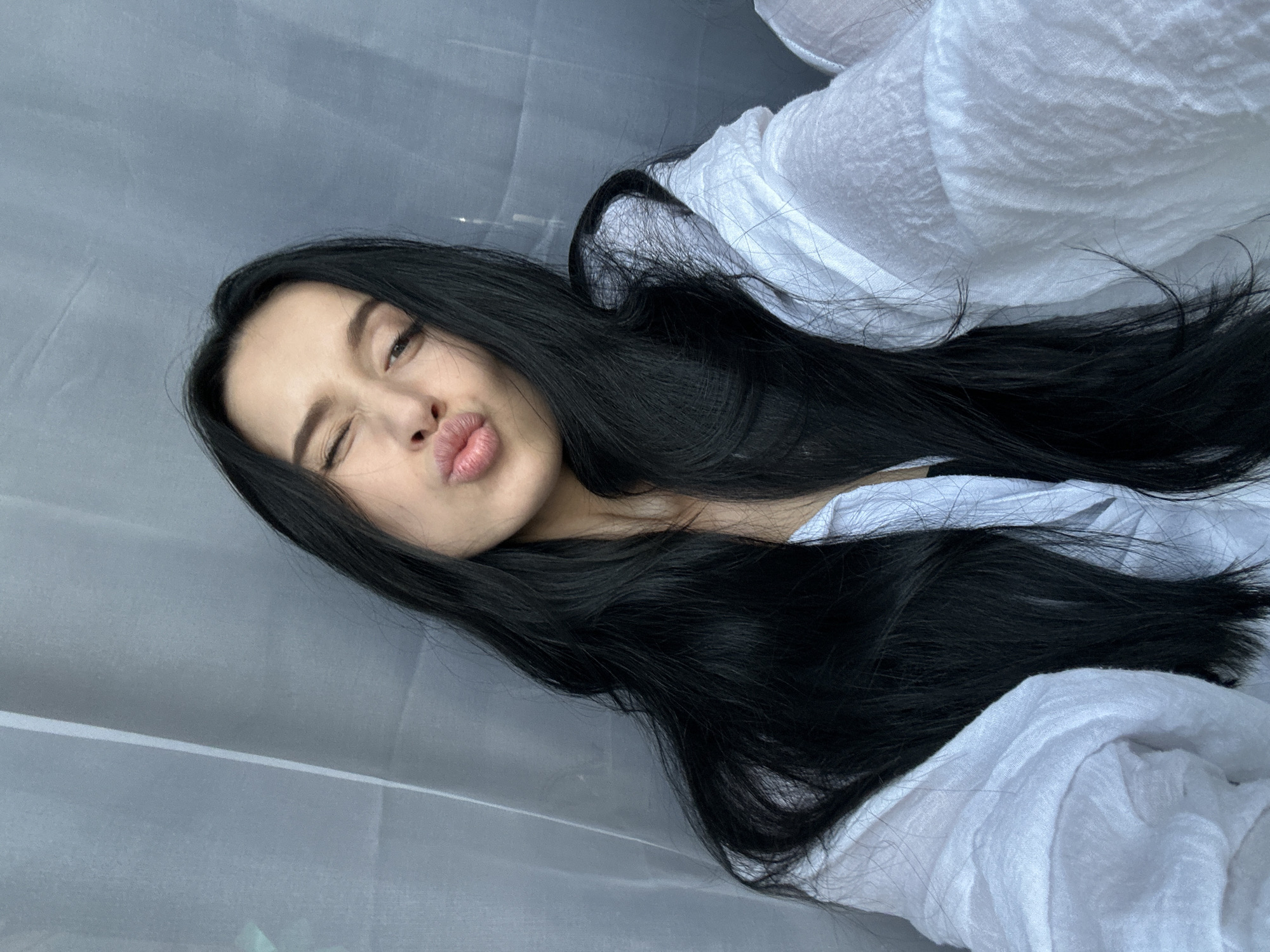 The image features a young woman with long, dark hair lying down. She is wearing a white garment, possibly a shirt or dress, made of a textured fabric. Her head is tilted, and she is looking towards the camera with one eye closed and her lips puckered in a playful expression, suggesting she is making a kissy face. The background is a soft, grey fabric, possibly a curtain or backdrop, with some indistinct texture. The lighting appears to be soft and diffused, casting gentle shadows. There are no visible signs of the time of day or specific weather conditions, and the location cues are limited to the indoor setting suggested by the fabric backdrop. The overall mood of the image is lighthearted and casual. There is no visible text in the image.
