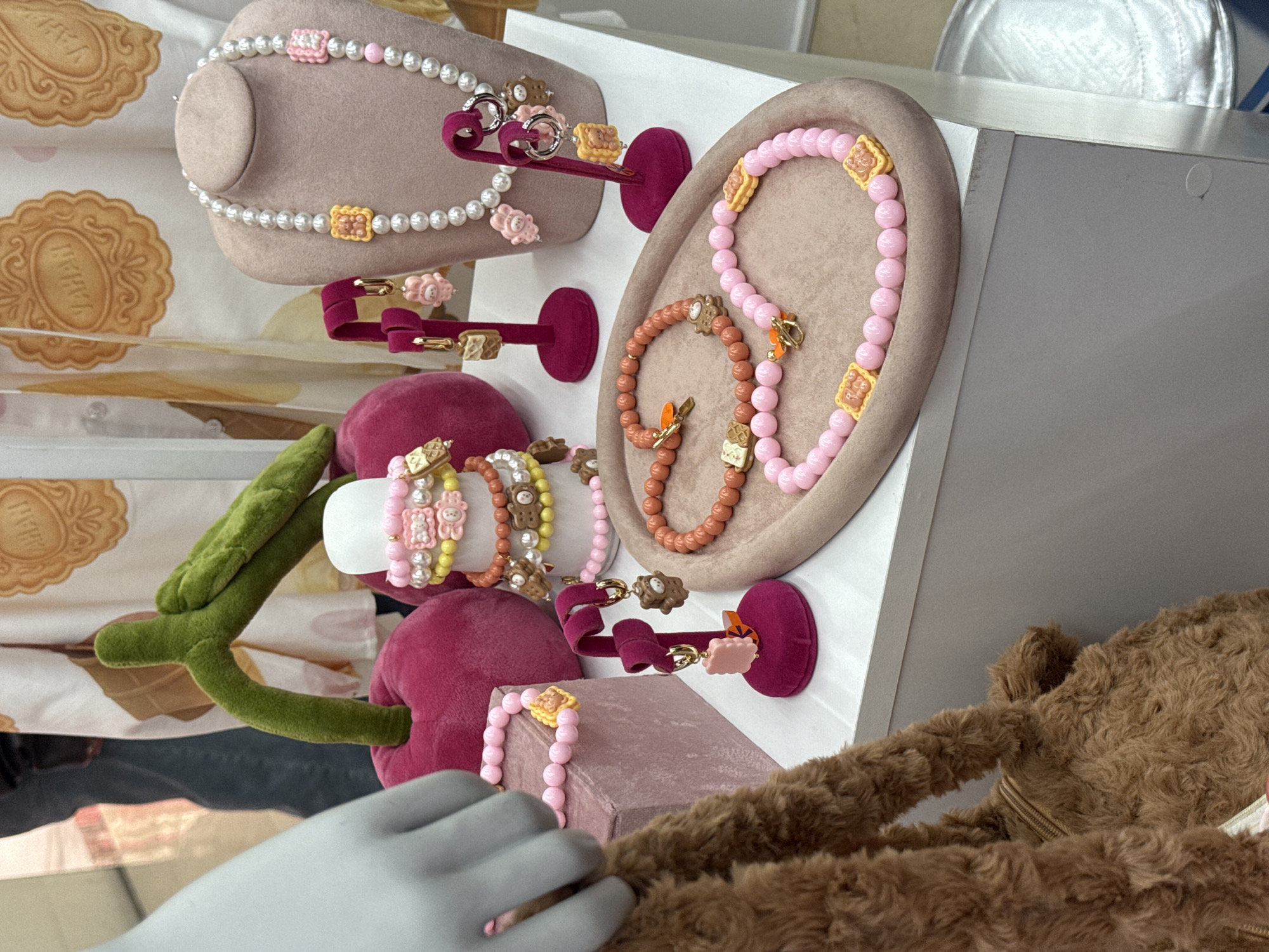 The image depicts a display of jewelry, including necklaces and bracelets, with various decorative elements such as teddy bear motifs and colorful beads. The display is arranged on multiple levels with mannequins showcasing the jewelry. A plush, brown teddy bear-like bag is also visible. The location is identified as Yerevan, Armenia.