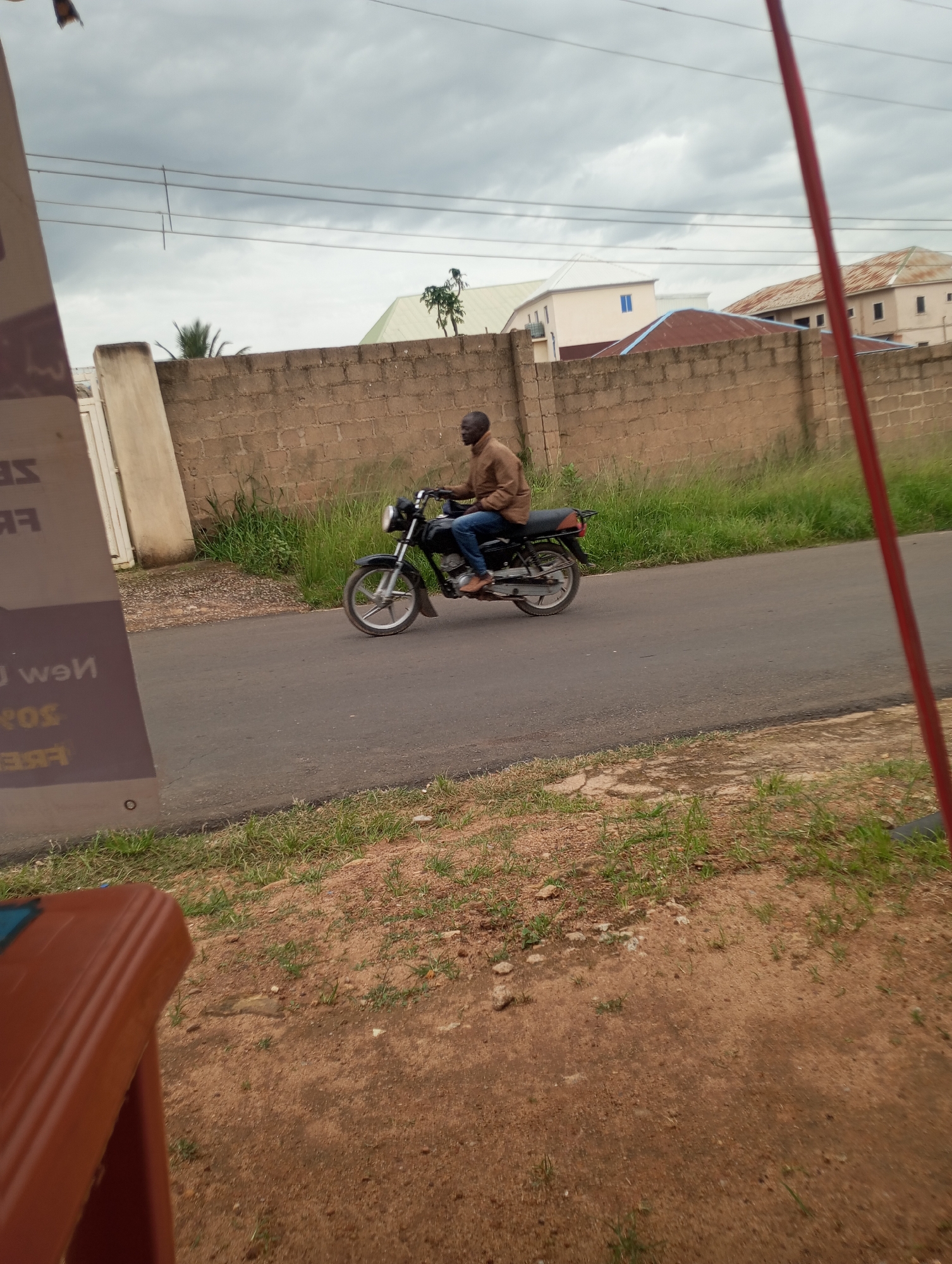 A man rides a black motorcycle on an asphalt road in Jos, Nigeria, under a cloudy sky. The man, wearing a brown long-sleeved shirt and blue jeans, is positioned in the center of the frame, with the motorcycle angled slightly towards the right. The road is bordered by a brick wall on the left, with overgrown green grass in front of it. Beyond the wall, several buildings with various roof colors are visible. In the foreground, a brown object, possibly a piece of furniture, partially obscures the lower left corner of the image. The overall environment suggests an urban or semi-urban setting.