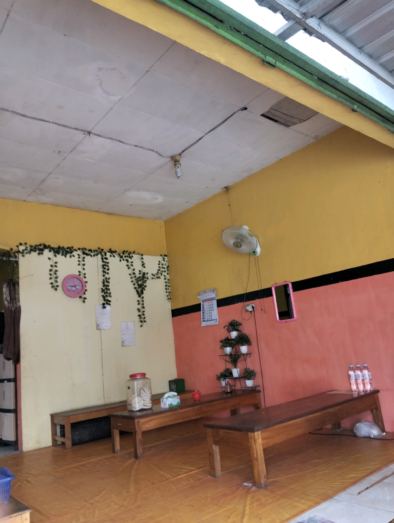 The image depicts an indoor seating area, possibly a small cafe or waiting room, in Ngawi, Indonesia. The space is characterized by its brightly colored walls, with the upper section painted yellow and the lower section a peachy-pink, divided by a black stripe. A portion of the ceiling shows damage, with a visible hole and water stains.
Wooden benches are arranged along the walls, suggesting a simple seating arrangement. On one table, a large glass jar filled with what appears to be crackers or snacks is prominent, next to a tissue box and a small green container. Another table holds a small red object, possibly a candle holder, and several bottles of water.
Decorative elements include artificial ivy draped along the yellow wall and a circular pink clock. A wall-mounted fan is visible in the upper right portion of the room, and a small pink-framed mirror hangs on the wall. A calendar, likely displaying dates for the current month, is also present. The floor is covered in a yellow mat. The overall impression is one of a utilitarian and somewhat rustic establishment.