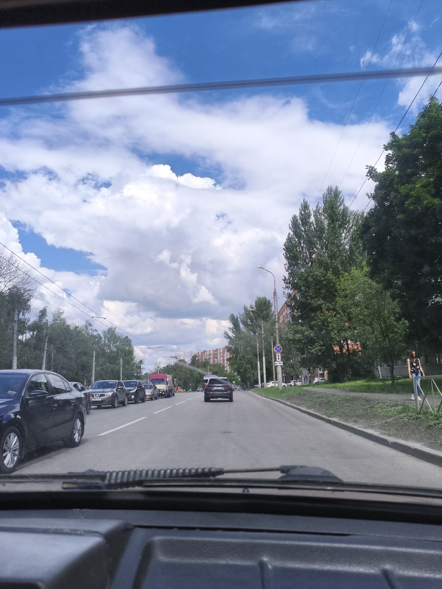 The image shows a view from inside a car looking out at a traffic scene on a cloudy day. The perspective is from the driver's seat, with the dashboard and windshield wipers visible in the foreground. The road ahead is lined with trees on the right and buildings in the distance. Several cars are in traffic, moving along the road. A woman is visible on the sidewalk on the right side of the road, walking away from the camera. The sky is blue with scattered white clouds. The scene appears to be in a residential area.