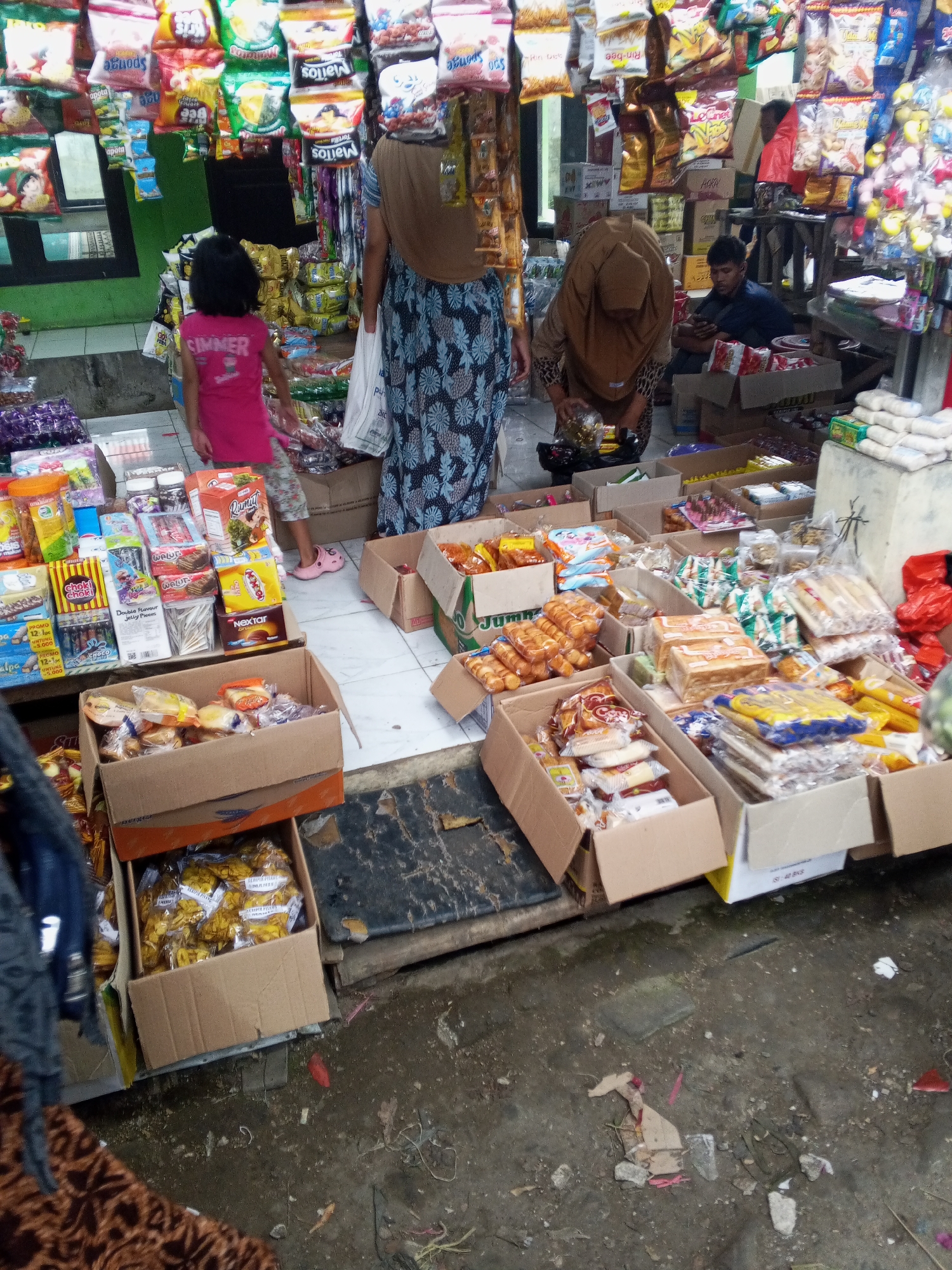 The image depicts an outdoor market stall in Serang, Indonesia. In the foreground, a young girl in a pink shirt and patterned shorts stands facing away from the camera, looking at a display of snacks. To her right, a woman wearing a brown headscarf and a patterned blue and white dress is seen from the side, bent over and interacting with items on the stall. Behind them, a man in a dark shirt is seated, looking at his phone.
The stall is laden with various packaged food items, predominantly snacks, arranged in cardboard boxes and hanging bags. The goods include chips, biscuits, and other packaged treats. The ground is dirt and appears somewhat wet and debris-strewn, indicating a typical market environment. In the background, more stalls and market activity are visible, with the overall impression being a busy, local marketplace. The lighting suggests it is daytime, possibly overcast or shaded.