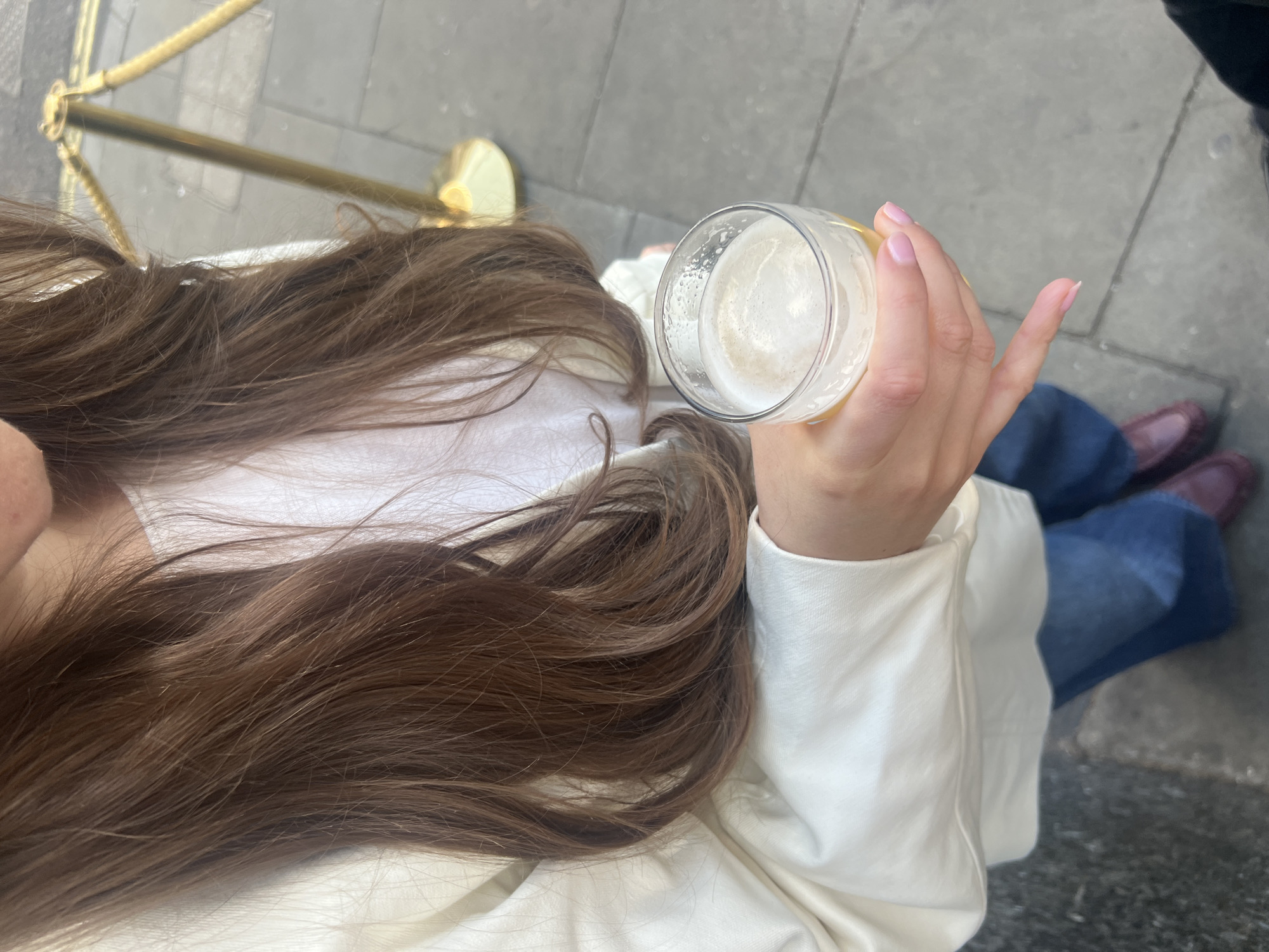 A high-angle shot captures a person holding a glass of beer. The person's long brown hair is prominently displayed. The person is wearing a white jacket and blue jeans. In the background are two reddish-brown shoes. The setting appears to be outdoors on a paved surface, possibly in a city, as suggested by the presence of a rope barrier, often found in public spaces. Without additional information, I cannot confirm that this photo was taken in London.
