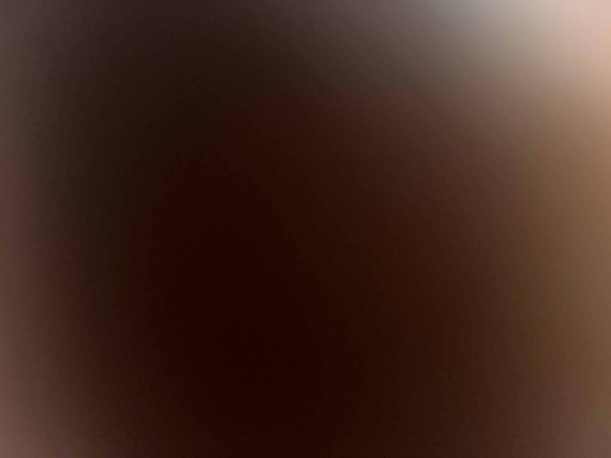 The image is a close-up, out-of-focus shot of a dark brown textured surface. The lighting appears to be dim, with a gradient of light from the top right corner that fades into the darker tones of the rest of the image. There are no discernible subjects, objects, or activities taking place. The texture of the surface is uniform, with a slight graininess visible. No text is present in the image. The overall impression is of a dark, abstract background.