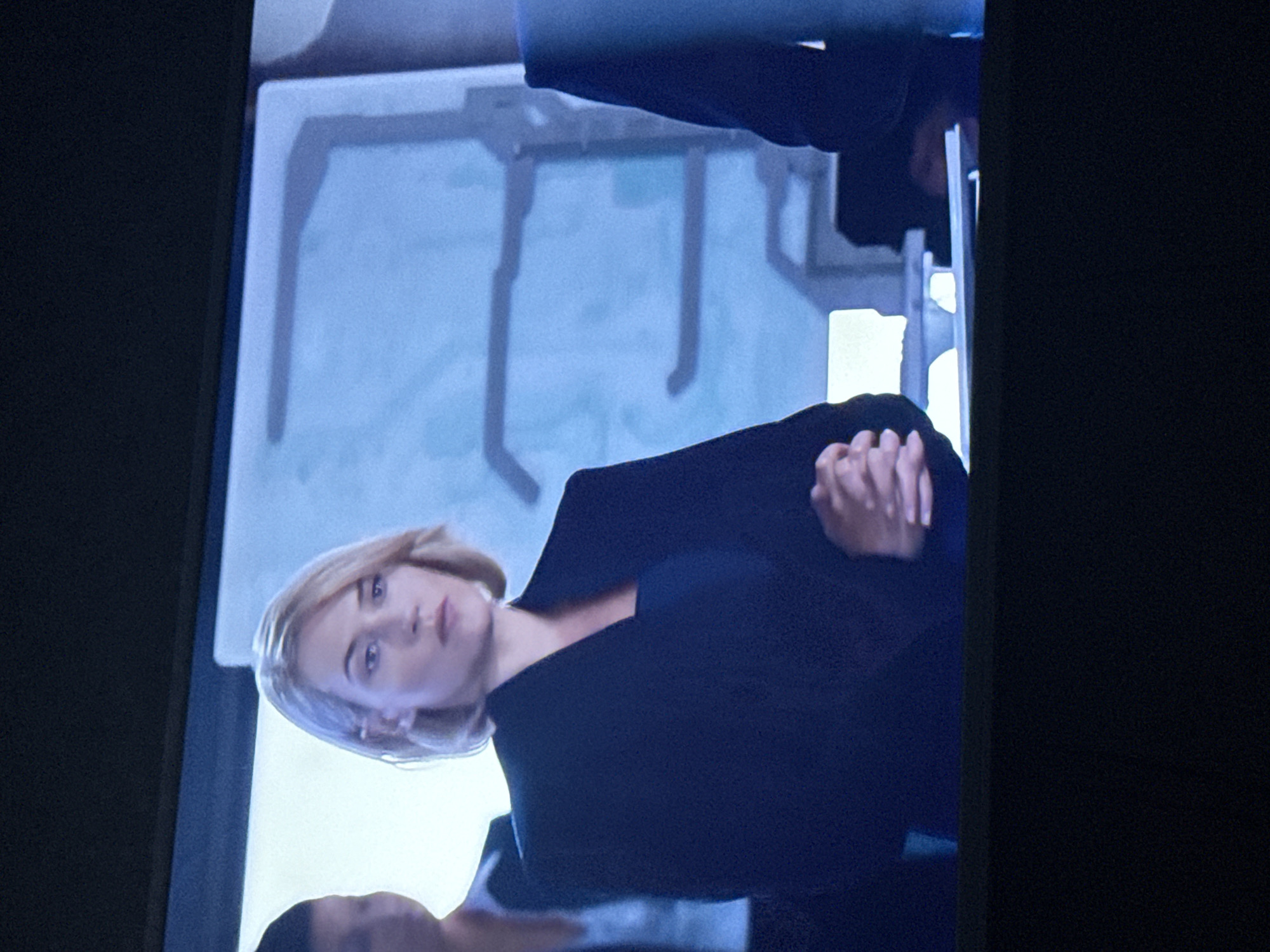 A medium shot of a woman with short blonde hair, wearing a dark-colored jacket or suit, standing with her hands clasped in front of her. There is a slightly out-of-focus background showing what appears to be a room or set with metallic structures. Another person's shoulder is visible in the bottom left corner. The image seems to be taken from a television screen. The location is not discernible from the visual information provided and therefore cannot be confirmed to be in Mytishchi, Russia.