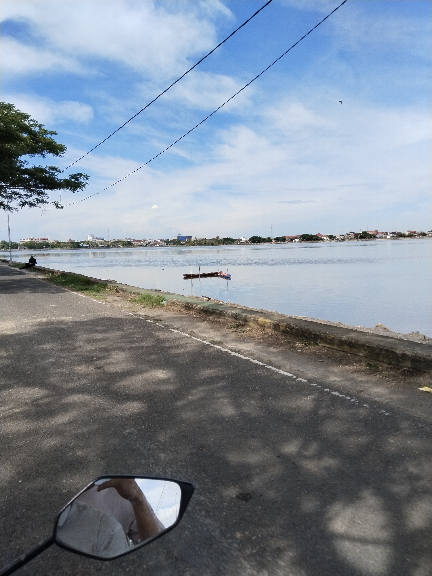 The image depicts a view of Waduk Lhokseumawe from a road alongside it. The road runs along the reservoir's edge, separated by a low concrete barrier with grass growing between the road and the barrier. The road appears to be asphalt and shows the shadows of trees. In the foreground, the rearview mirror of a motorcycle is visible, reflecting a person's arm and shirt. A person is sitting on the side of the road near a tree. In the reservoir, a small, moored boat is visible. Across the reservoir, there are buildings and trees, part of the urban landscape of Lhokseumawe. Overhead are power lines, and a bird is flying in the blue sky with scattered white clouds.
