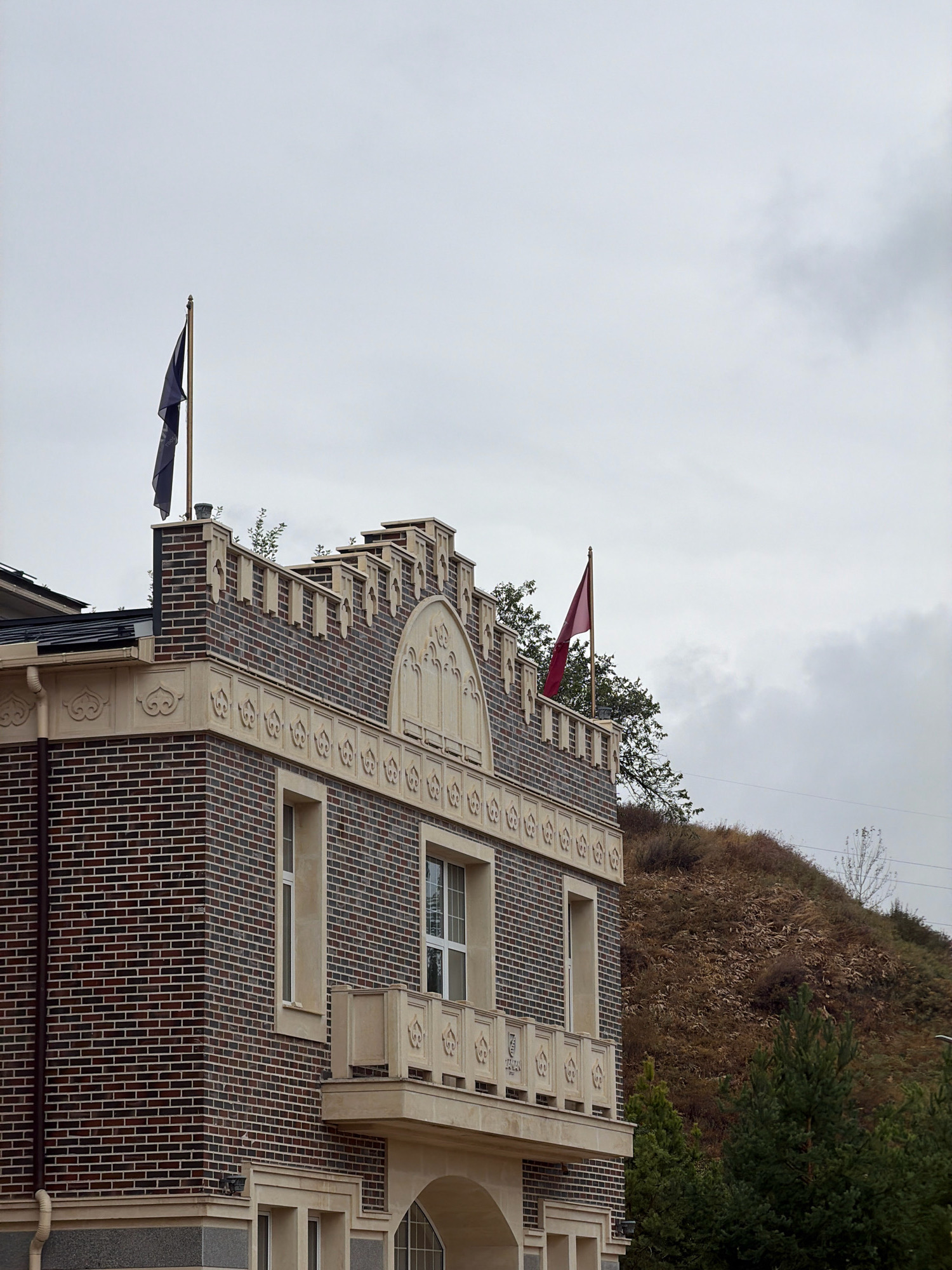A building with a castellated parapet and two flags flying from flagpoles is shown against a cloudy sky. The building's facade is constructed of dark brick with light-colored decorative elements and a stone base. A balcony with decorative railings extends from the second story. To the right, a hillside covered in dry vegetation rises behind the building, with evergreen trees in the foreground. Power lines are visible against the sky. The building appears to be a modern structure designed with historical architectural influences. The location is identified as Almaty, Kazakhstan.
