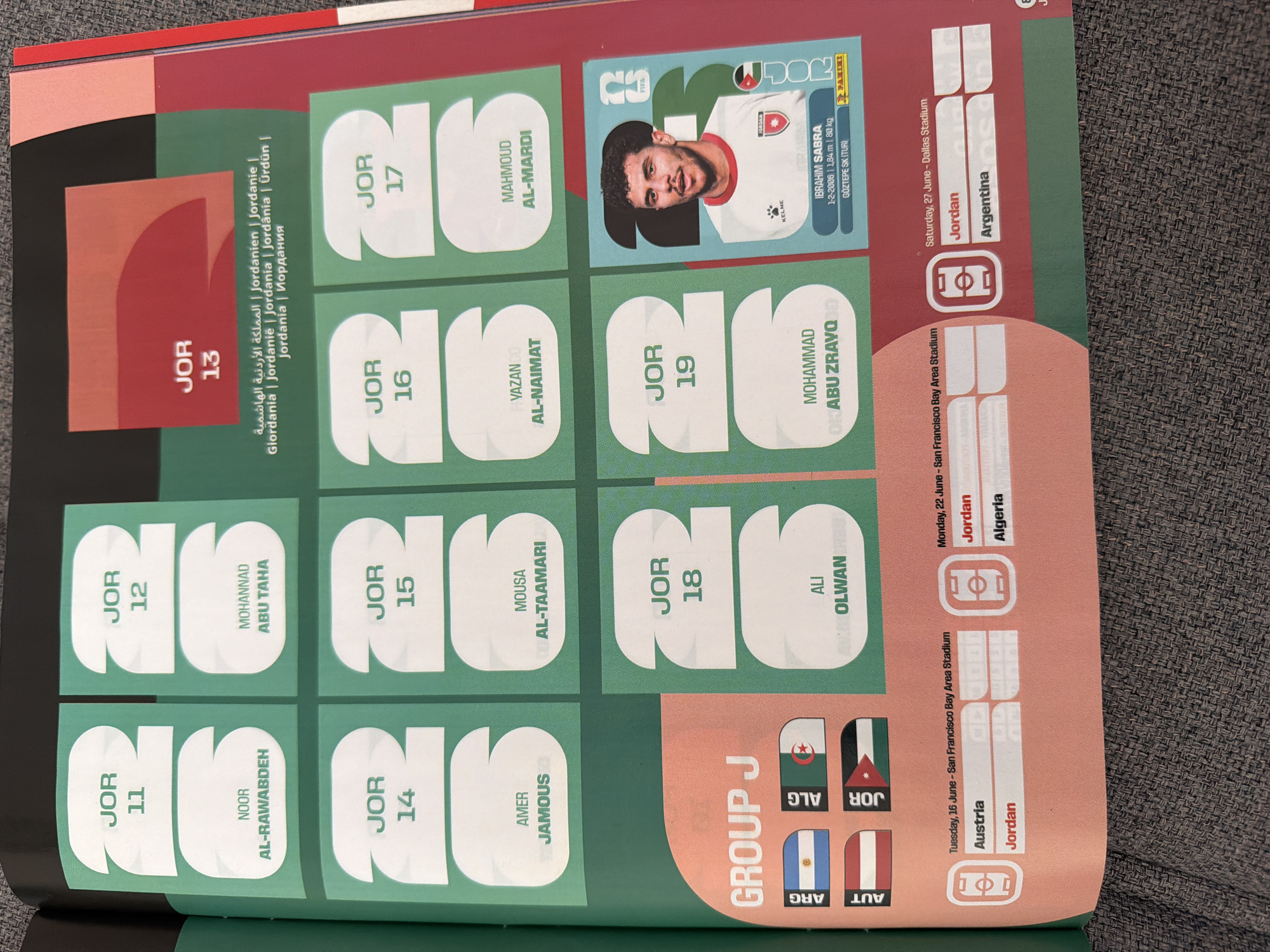 The image shows a page from a sticker album, likely related to a football (soccer) tournament. The page displays stickers of football players from Jordan, indicated by "JOR" on each sticker. The players' names and jersey numbers are visible, such as "JOR 11 NOOR AL-RAWABDEH", "JOR 12 MOHANNAD ABU TAHA", "JOR 13", "JOR 14 AMER JAMOUS", "JOR 15 MOUSA AL-TAAMARI", "JOR 16 VAZAN AL-NAIMAT", "JOR 17 MAHMOUD AL-MARDI", "JOR 18 ALI OLWAN", and "JOR 19 MOHAMMAD ABU ZRAVQD". There is also a larger sticker of player IBRAHIM SABRA with his details. Additionally, the page shows fixtures for "GROUP J" with opponent flags and match dates and locations, including "San Francisco Bay Area Stadium" and "Dallas Stadium". There are no indications of the city of Zlatitrap, Bulgaria, in the image. The overall environment appears to be indoors, with the page laid on a textured surface, possibly fabric.