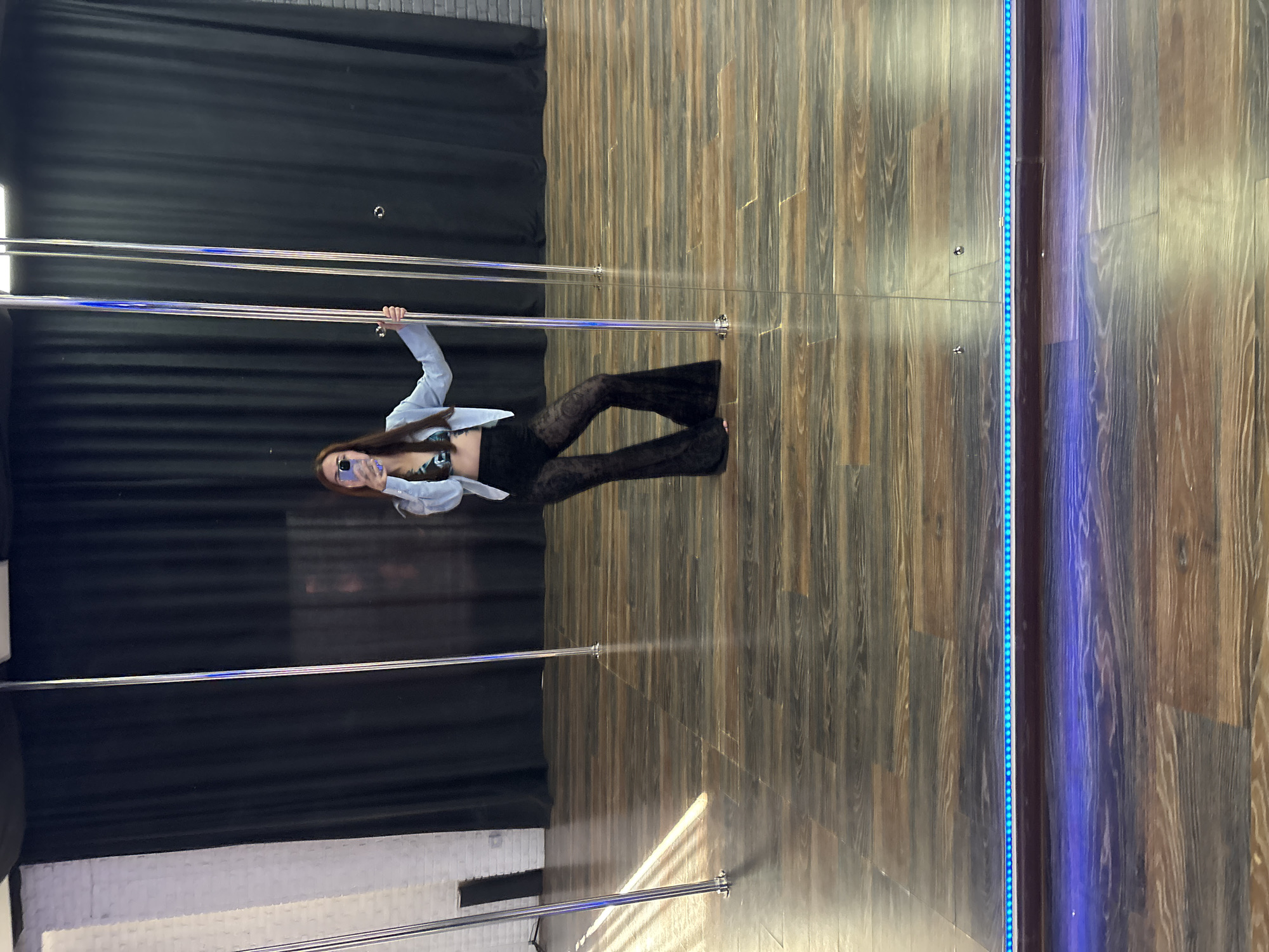 A person is holding onto a pole in what appears to be a dance studio or gym. The person is wearing a light blue jacket over a dark-colored top and black flared lace pants. They are also wearing glasses with purple lenses and are holding up a phone, seemingly to take a picture. The floor is made of wooden planks, and there is a black curtain in the background. To the right of the pole, there is a glass panel with blue LED lights running vertically along its edge. The lighting suggests it might be daytime, though the specific time is unclear. The scene is indoors, and there are no indicators of weather conditions. The overall mood seems to be relaxed and perhaps focused on practice or personal expression. The location is identified as Naberezhnye Chelny, Russia.
